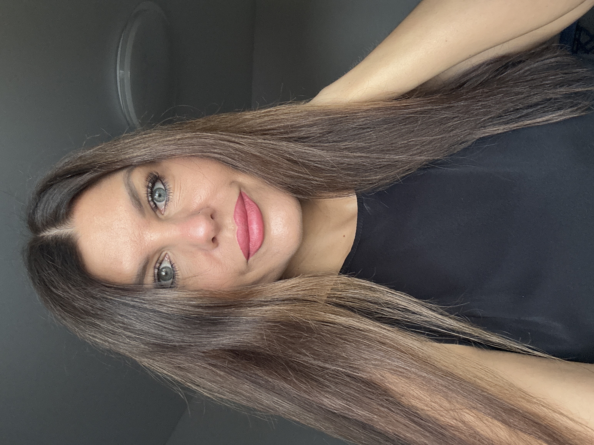 A medium shot, head and shoulders, features a woman with long, straight, brown hair with lighter streaks. She is looking directly at the camera, with her left hand touching the back of her neck. The woman has light blue eyes, prominent eyelashes, and light pink lipstick. She is wearing a black sleeveless top. The background is a plain, dark gray wall with a circular light fixture visible in the upper right portion of the frame. There is no indication of the city of Chaikovsky, Russia, in the image.