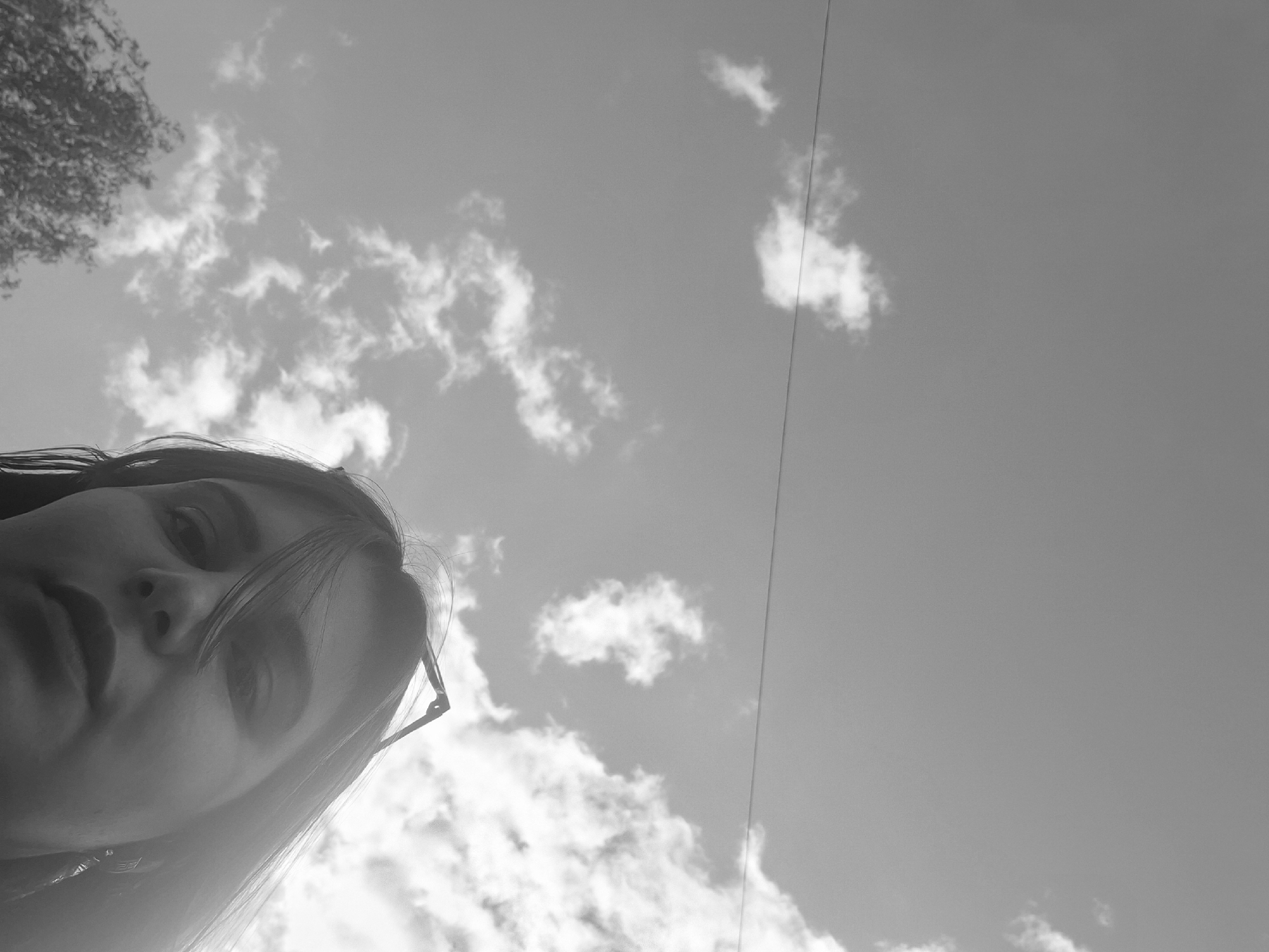 A grayscale, low-angle selfie depicts a young woman with fair skin and dark hair wearing glasses. She is positioned in the lower-left quadrant of the frame. Her face is angled towards the sky, occupying a significant portion of the view. The background features a partly cloudy sky. A power line runs diagonally across the sky. Some tree foliage is visible in the upper-left corner. The location is outdoors, potentially in Chaikovsky, Russia.