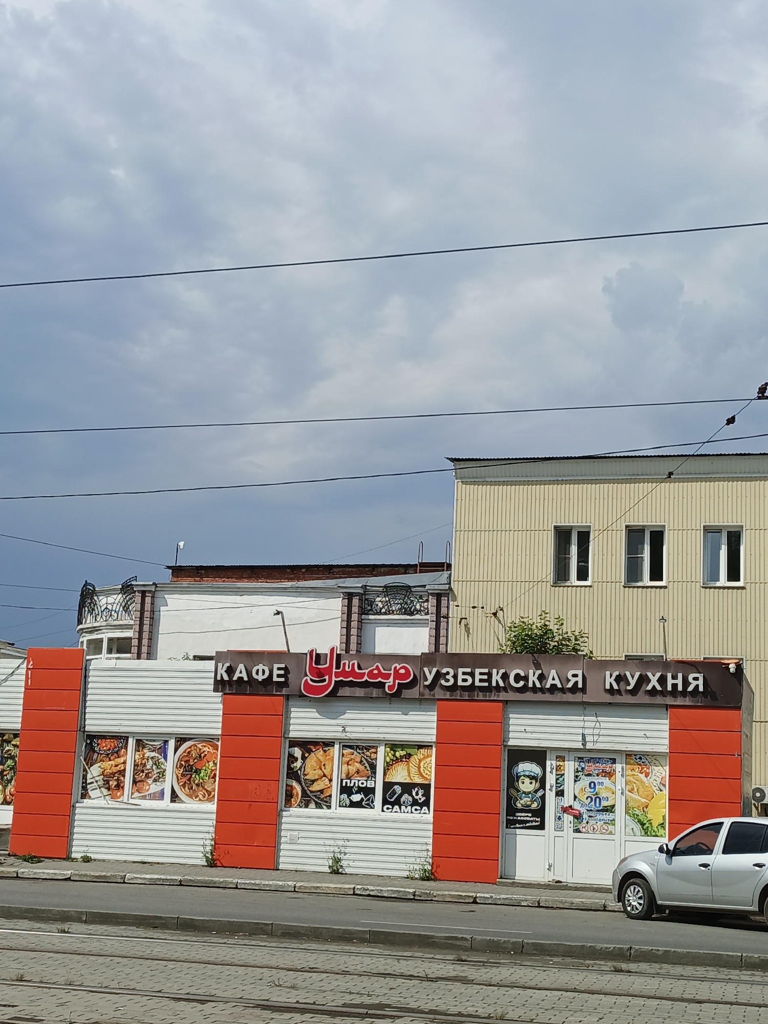 This is a photograph of a cafe named "Умар" (Umar) with "УЗБЕКСКАЯ КУХНЯ" (Uzbek Cuisine) written above it, located on a city street in Zlatoust, Russia. The cafe has a white corrugated metal facade with bright orange vertical accents. Several large windows display images of Uzbek dishes, including pilaf and samsa. A silver car is parked in front of the cafe. The sky above is cloudy. The scene appears to be a commercial area with overhead power lines visible.