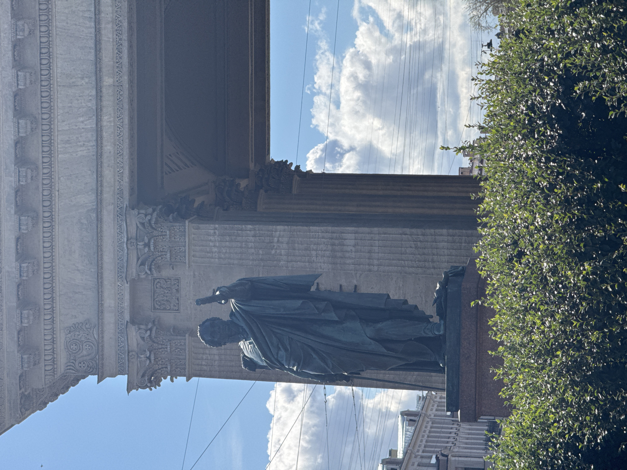 The image depicts the Kazan Cathedral in Saint Petersburg, Russia. In the foreground, a dense green hedge partially obscures the view. Behind the hedge, a large bronze statue of General Mikhail Kutuzov stands on a granite pedestal, his right arm raised and holding a baton. The statue is set against the backdrop of the cathedral's colonnade, a monumental semicircular structure with massive Corinthian columns. The sky is blue with scattered white clouds, and power lines are visible crisscrossing the frame. The architecture suggests a sunny day in the city.