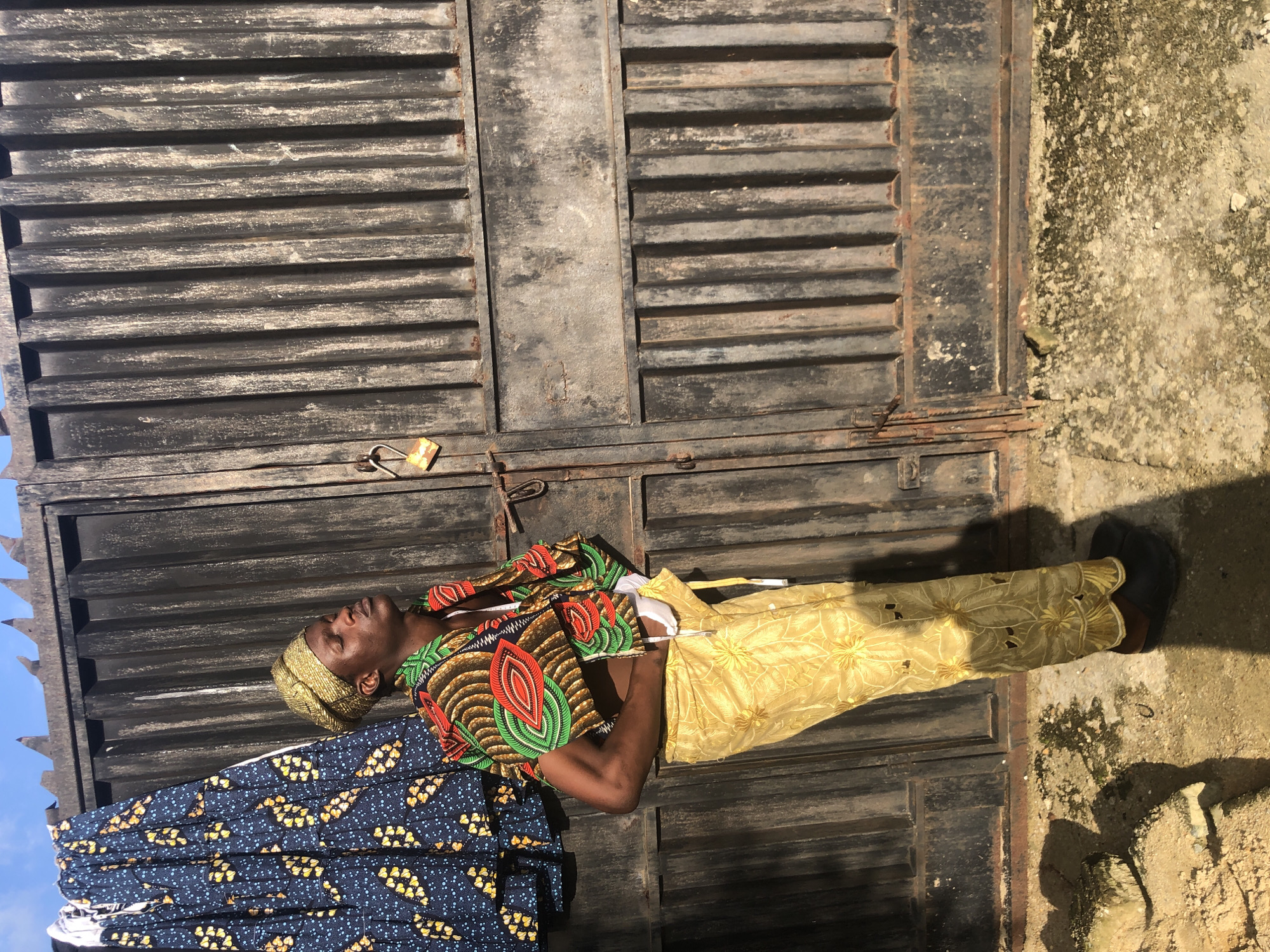 A man stands in front of a corrugated metal gate in Ado, Nigeria, wearing a gold-colored headwrap, a colorful patterned short jacket, and gold patterned pants. He has his eyes closed and head tilted back. To the left of the man, a patterned blue and yellow cloth hangs from a clothesline. The gate has a rusted padlock attached. The ground is a mix of concrete and dirt. The scene is illuminated by bright sunlight, casting distinct shadows.