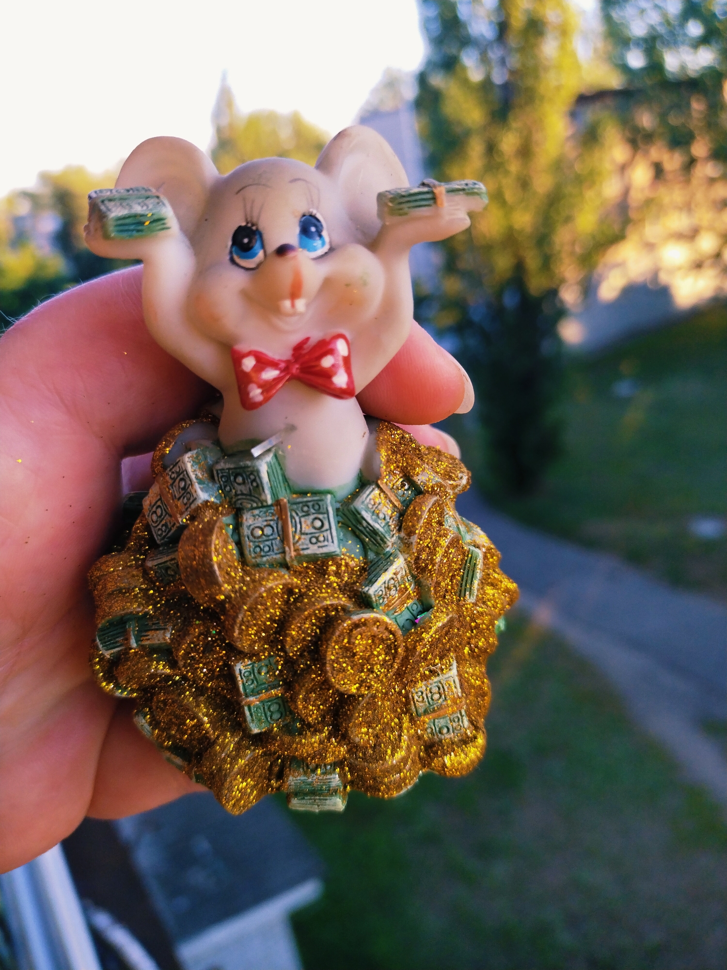 A close-up, eye-level, outdoor shot features a figurine of a light-skinned, cartoonish mouse held by a person's hand against a blurred background of trees and a path. The mouse is wearing a red bow tie with white polka dots. Its skirt is made of what appear to be small, stacked bundles of imitation money, some of which are green and some of which are gold and glittery. The mouse is holding two small bundles of what look like money in its outstretched hands. The person's thumb and index finger are visible on the left side of the frame, supporting the figurine. The lighting suggests it is either sunrise or sunset, casting a warm glow on the scene. The background is softly focused, indicating a shallow depth of field, and includes greenery and a paved walkway. The specific location in Horishni Plavni, Ukraine cannot be determined from the visual information in the image.