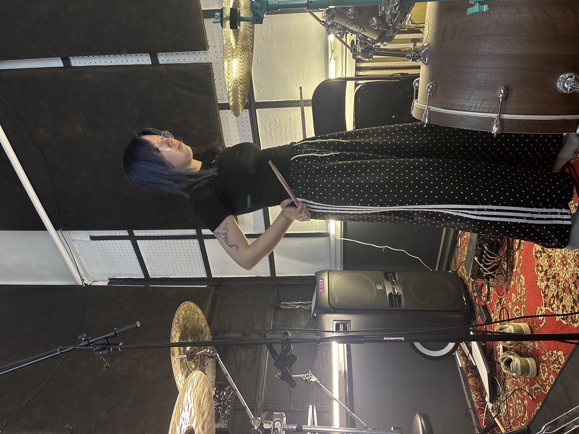 In a recording studio, a young person with blue-dyed hair and glasses stands near a drum set, holding drumsticks. They are wearing a black t-shirt and black pants with white polka dots and white stripes on the sides. The studio has soundproofing panels on the walls and a patterned rug on the floor. A large speaker and audio equipment are visible. The person is positioned between a microphone stand and the drum set, suggesting they are about to play or have just finished playing. There are no other people or animals in the frame. The scene is well-lit, with the primary light source appearing to be overhead. The location is identified as Chaikovsky, Russia.