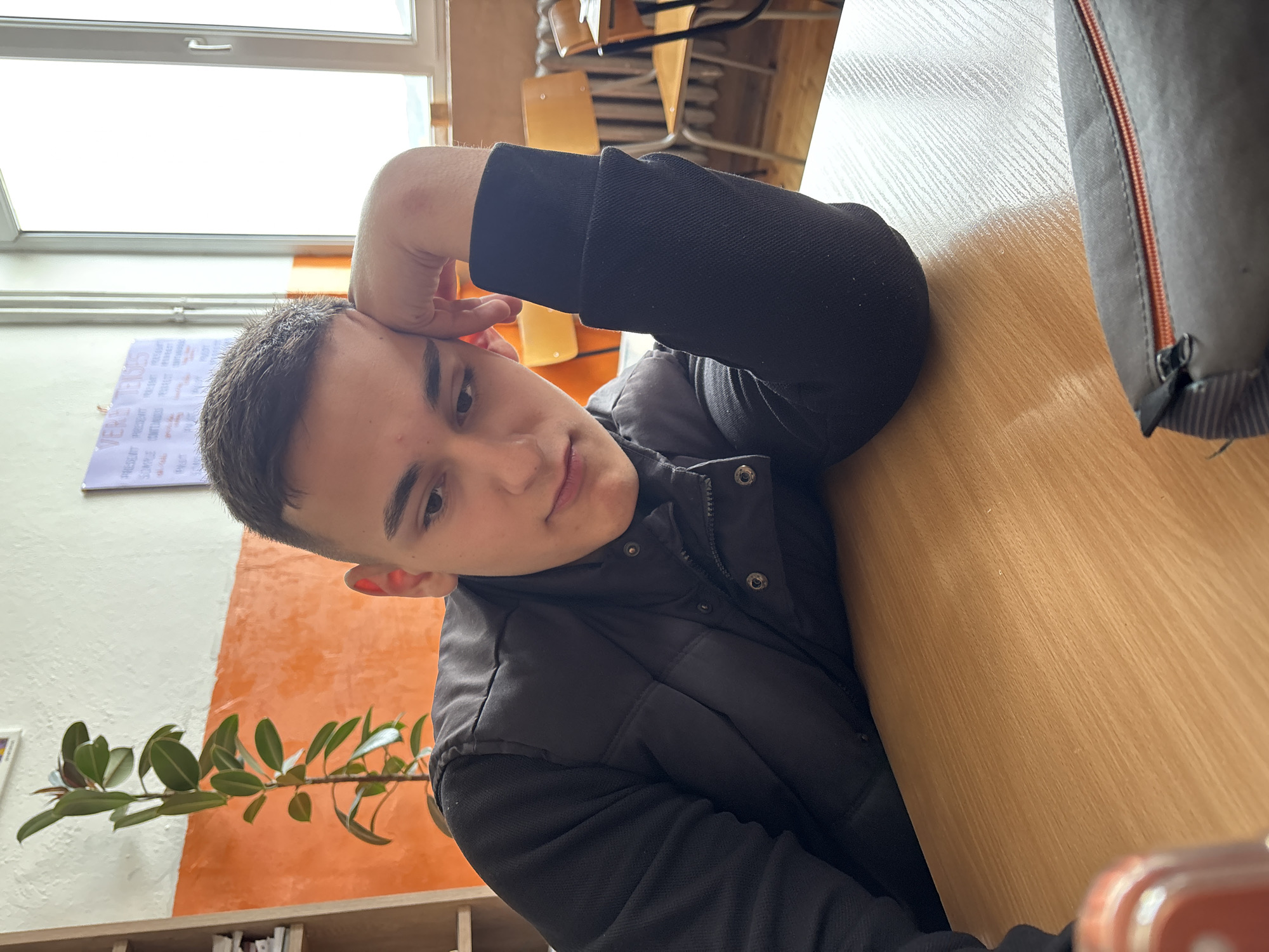 A young male with short dark hair and a neutral expression is captured in a portrait shot, looking directly at the camera. He is wearing a black long-sleeved top and a black puffer vest, with his left hand resting on his forehead.
The setting appears to be an indoor educational environment, likely a classroom or study area in Panagyurishte, Bulgaria. A light-colored wooden desk occupies the foreground and right side of the image, with a gray bag featuring an orange zipper partially visible on its surface. In the background, a large white-framed window allows bright daylight into the room, suggesting it is daytime. On the wall to the left, there is a poster or chart with French verb conjugations visible, including the prominent word "VERBES" and categories like "PRÉSENT," "PASSÉ COMPOSÉ," "IMPARFAIT," and "FUTUR SIMPLE." The wall itself is painted white on top and a contrasting orange below, with a green leafy plant positioned in front of the orange section. Behind the person and visible through the window, stacks of wooden chairs and desks further reinforce the classroom setting.