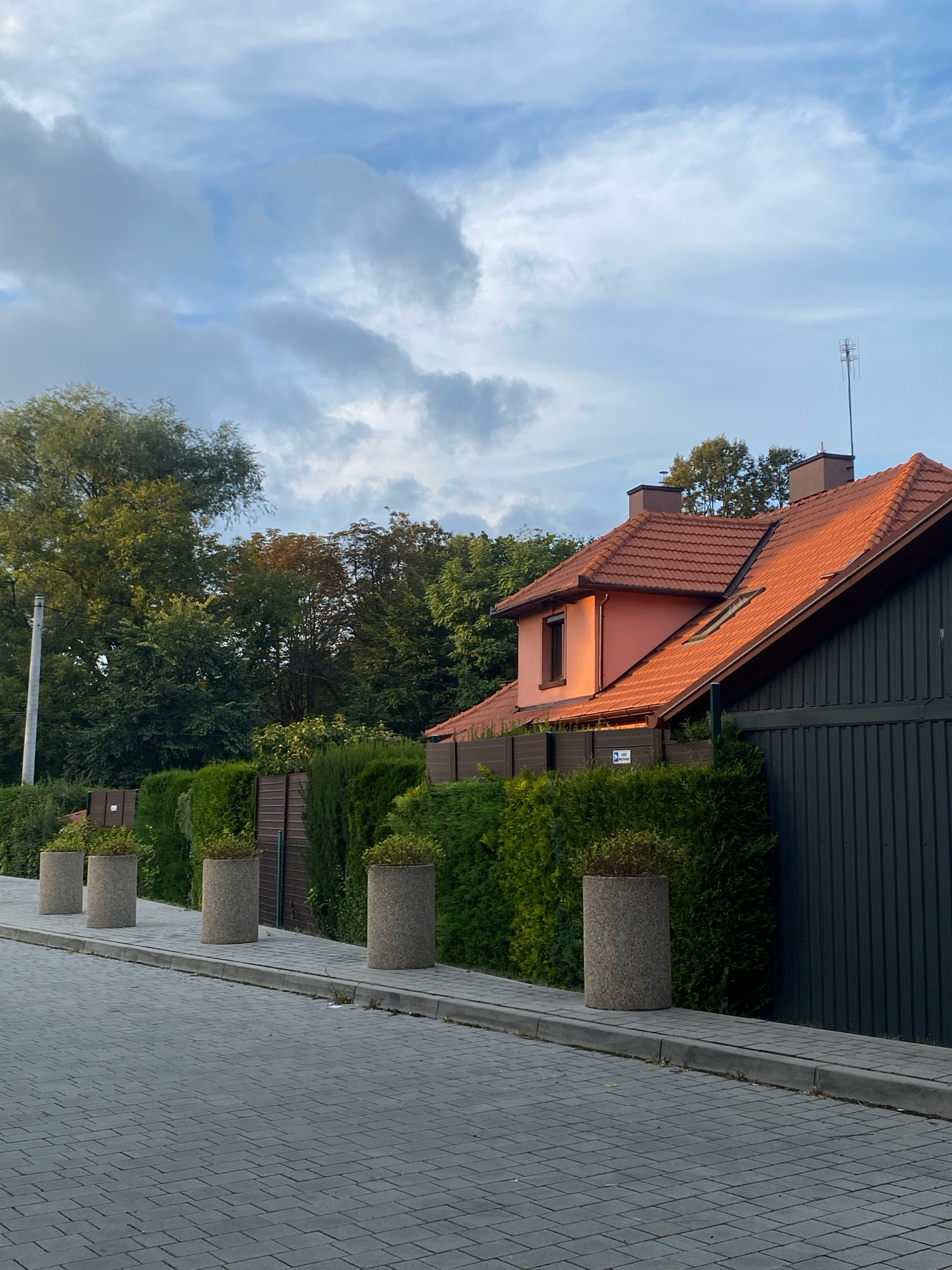 The image shows a residential street in Nowy Sącz, Poland, with a house on the right side and a sidewalk lined with planters and hedges. The house has a red tile roof and a light pink wall, with a window illuminated by the setting sun. A dark fence is visible behind the hedges, and a television antenna is mounted on the roof. The street is paved with gray interlocking bricks, and the sidewalk is made of concrete slabs. The sky is partly cloudy, with a mix of blue and white clouds. The scene appears to be set in the late afternoon or early evening, as indicated by the warm lighting on the house. There are no people or animals visible in the image. The overall impression is one of a quiet, well-maintained suburban neighborhood.
