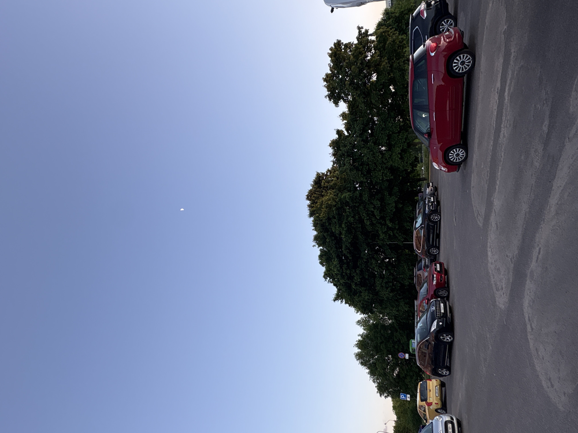 The image shows a parking lot in Minsk, Belarus. Several cars are parked in a row, including a red Fiat 500L, a black car, and a yellow car. Trees line the parking lot. The sky is blue with a crescent moon visible.