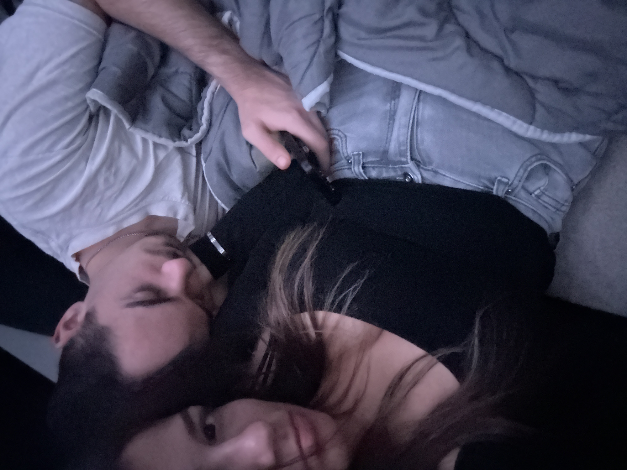 A couple is lying down, possibly in a bed or on a couch, under a gray comforter. The man is wearing a white t-shirt and the woman is wearing a black shirt. The man's left arm is over the comforter. The location of the image is unknown and cannot be determined to be in Sofia, Bulgaria.