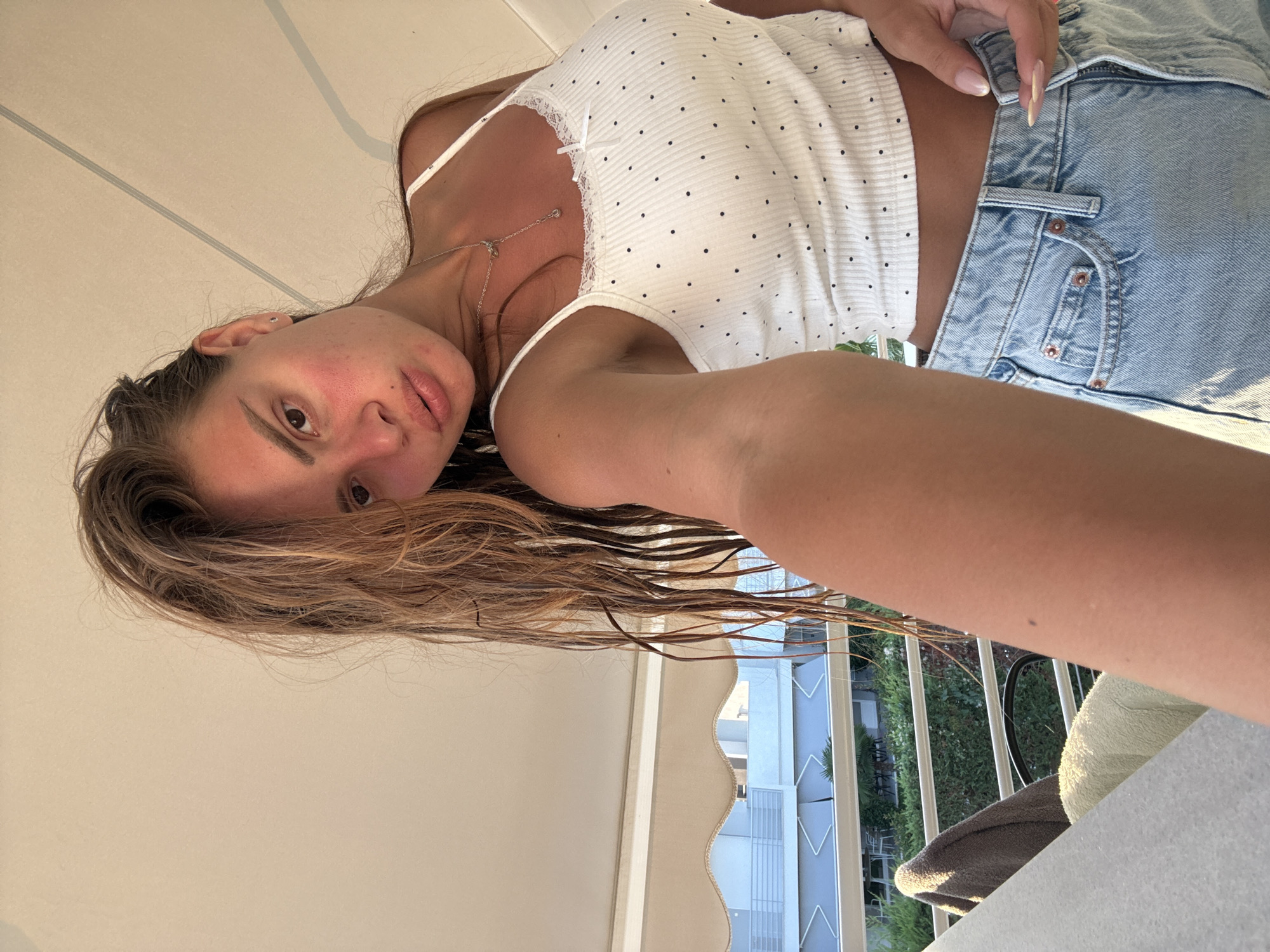 A young Caucasian woman with long, wet, light brown hair takes a selfie on a balcony overlooking a building and green foliage. She is wearing a white, polka-dot camisole and light blue denim shorts. Her face shows a flushed appearance on her cheeks. The background includes a beige awning and railings.