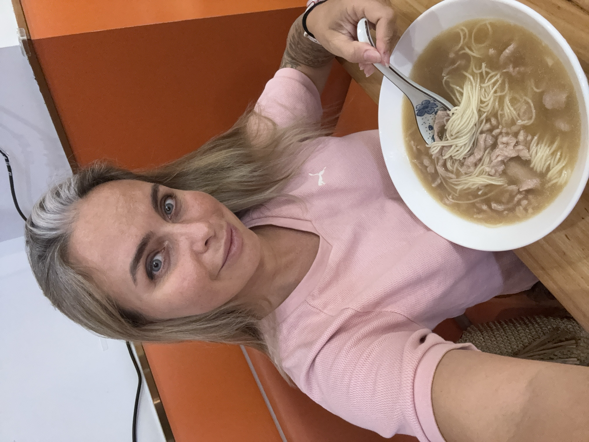 A selfie captures a woman with blonde hair, streaks of gray, and blue eyes. She is wearing a pink collared shirt with a Puma logo. She is seated at a wooden table in what appears to be a casual dining establishment, possibly a noodle shop, with orange booth seating in the background. She is holding a spoon with a blue floral pattern, lifting noodles from a bowl of soup containing noodles and thinly sliced meat. The environment suggests a locale within Shanghai, China, given the commonality of such eateries and the typical interior design choices found in the city.