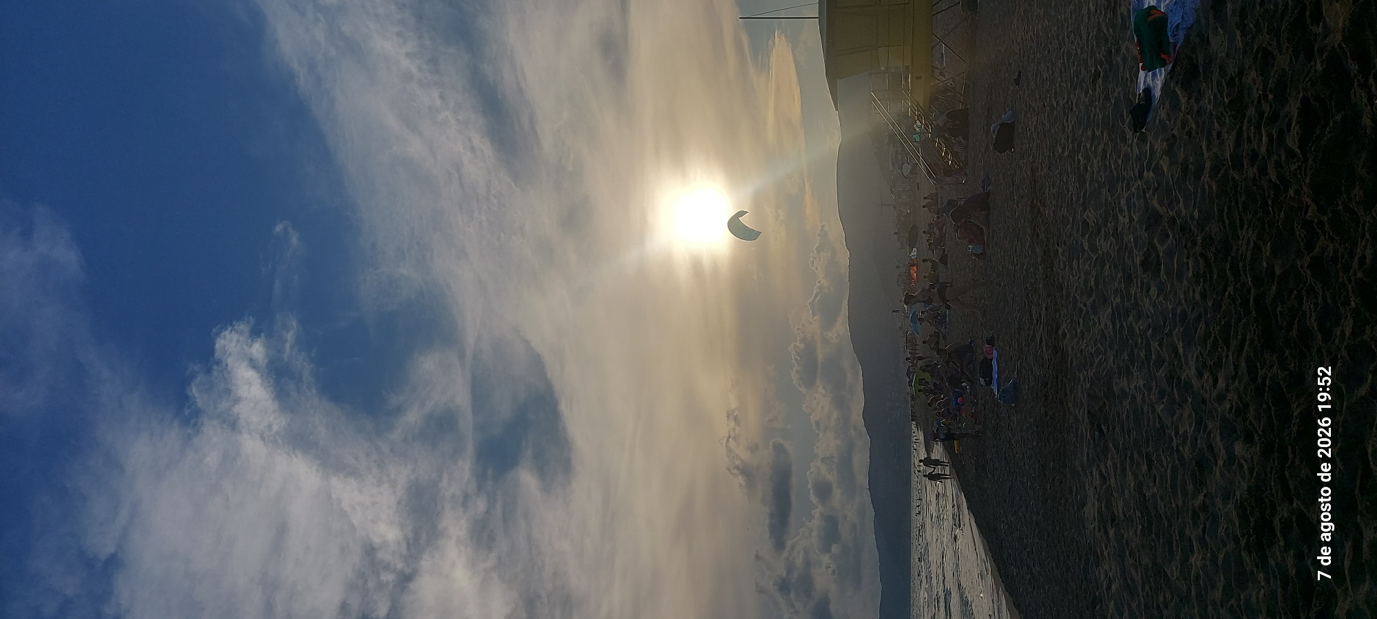 The image depicts a crowded beach scene at sunset, likely in Castelldefels, Spain, as indicated by the timestamp "7 de agosto de 2026 19:52". The sun is low in the sky, casting a warm glow and creating lens flare. In the upper portion of the image, a kite surfer is visible, soaring against the bright sun. The beach itself is populated with numerous people, many of whom are seated or lounging on the sand. There are umbrellas providing shade, and a yellow lifeguard tower stands prominently on the right side of the frame. The sea is visible on the left, with gentle waves lapping the shore. In the background, hills or mountains form a silhouette against the twilight sky. The sand appears dark, suggesting it might be volcanic or wet. The overall atmosphere is one of leisure and recreation on a summer evening.