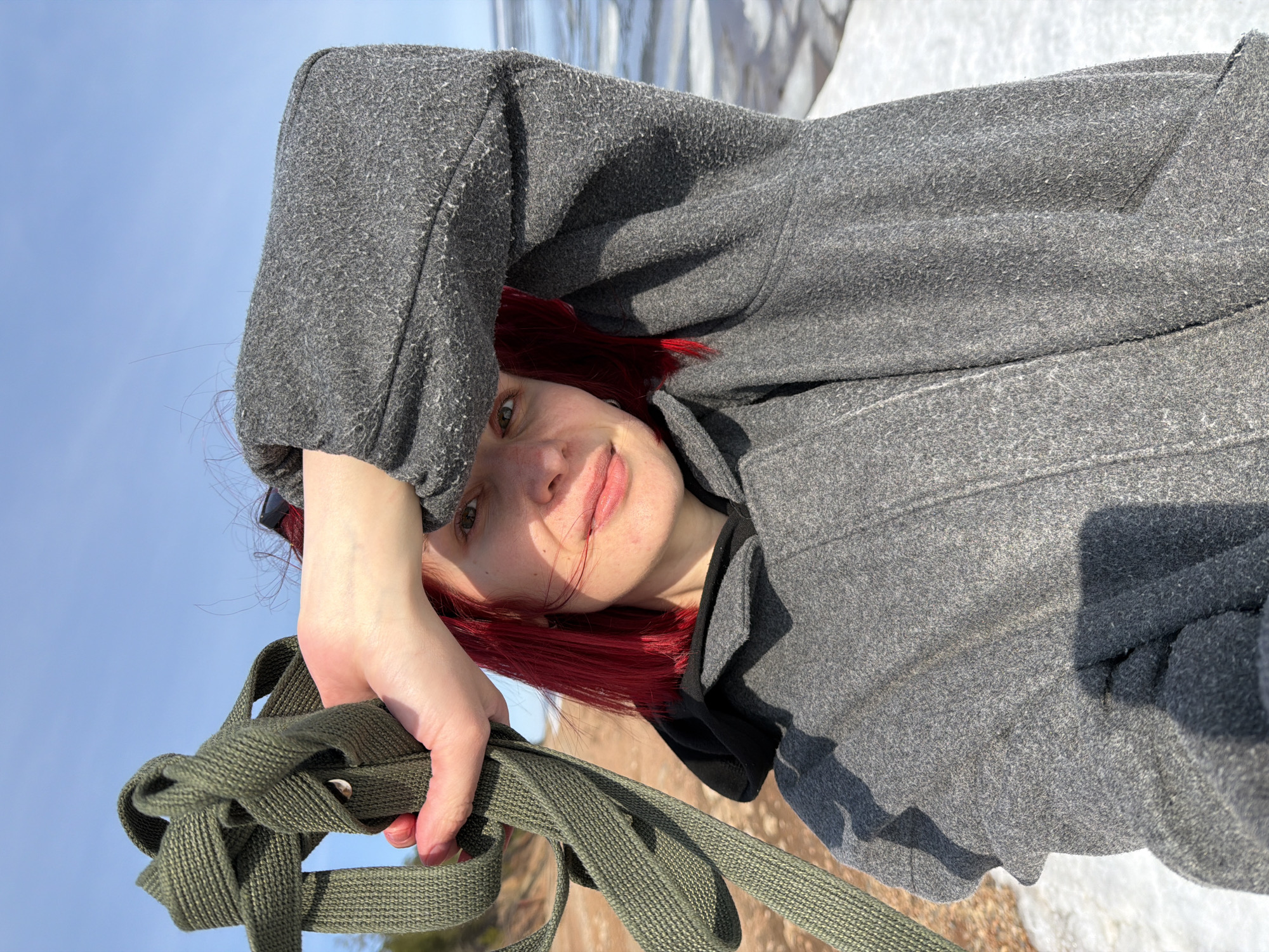 A person with red hair wearing a gray coat is holding a knotted olive green strap. The background shows a patch of snow, with a landscape of brown and white. The sky is blue. The location is Noviy, Russia.