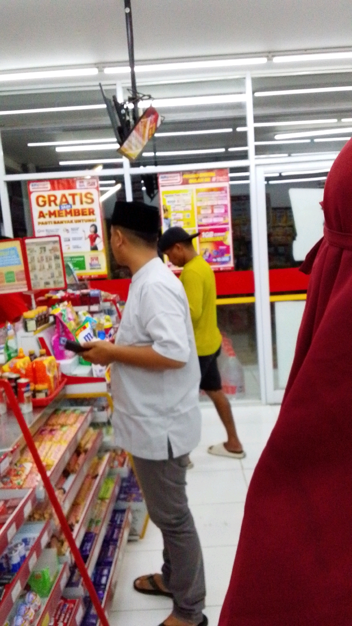 The image shows the interior of a convenience store, possibly an Alfamart or Indomaret, in Desa Karangbenda, Indonesia. In the foreground, a man wearing a white koko shirt, grey trousers, and a black peci cap is holding a black object, possibly a scanner or a phone. He is facing away from the camera and looking at a display of goods on shelves. To his right, another man in a yellow t-shirt and dark shorts is partially visible, also looking at the shelves. A portion of a person wearing a maroon hijab is visible on the far right edge of the frame. The shelves are stocked with various food items, snacks, and other merchandise. In the background, promotional posters are displayed on the walls. The lighting suggests it is daytime inside the store.