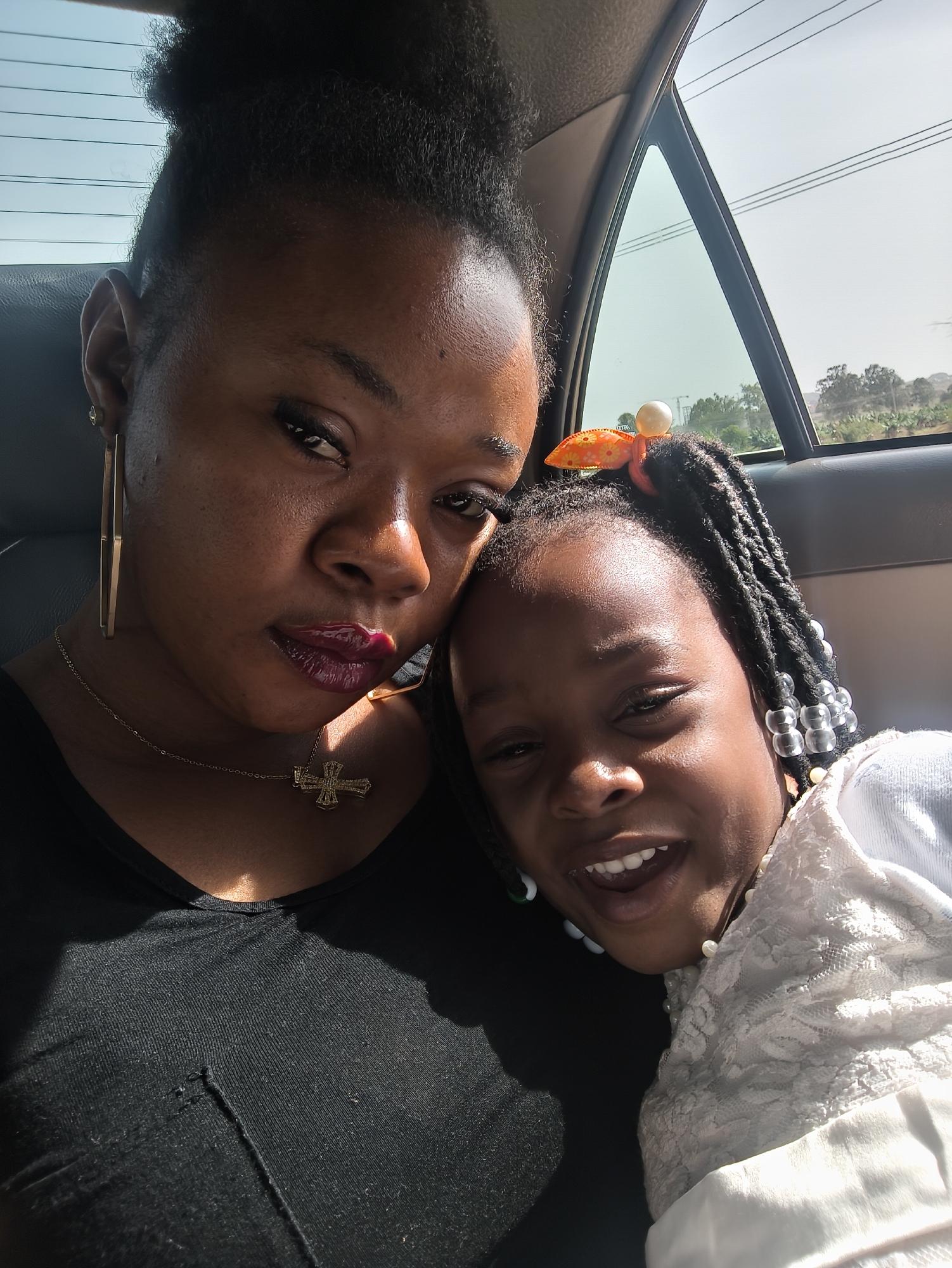This close-up portrait captures an adult woman and a young girl, likely a mother and daughter, inside a car in Jos, Nigeria. The adult woman, positioned on the left, has dark skin and is looking directly at the camera with a contemplative expression. She wears a black V-neck top, large gold-toned rectangular hoop earrings, and a gold-colored necklace with a prominent cross pendant. Her dark hair is styled up, and her lips are adorned with glossy, dark red lipstick.
To her right, nestled close, is a young girl with dark skin, smiling broadly with an open mouth, revealing her teeth. She wears a white top, possibly with a lace overlay. Her hair is braided, decorated with clear beads, and she sports an orange patterned hair clip featuring a white, pearl-like ornament.
The scene is brightly lit by natural light, suggesting it is daytime. Strong highlights fall across their faces, particularly on the woman's forehead and nose, and the girl's cheeks. Through the car window in the background, power lines stretch across a bright, clear sky. Below the power lines, distant green foliage, possibly indicative of banana trees, and a construction crane are visible, hinting at a semi-urban or rural landscape. The tender embrace and joyful expression of the child convey a warm, affectionate interaction between the subjects. No text is visible in the image.