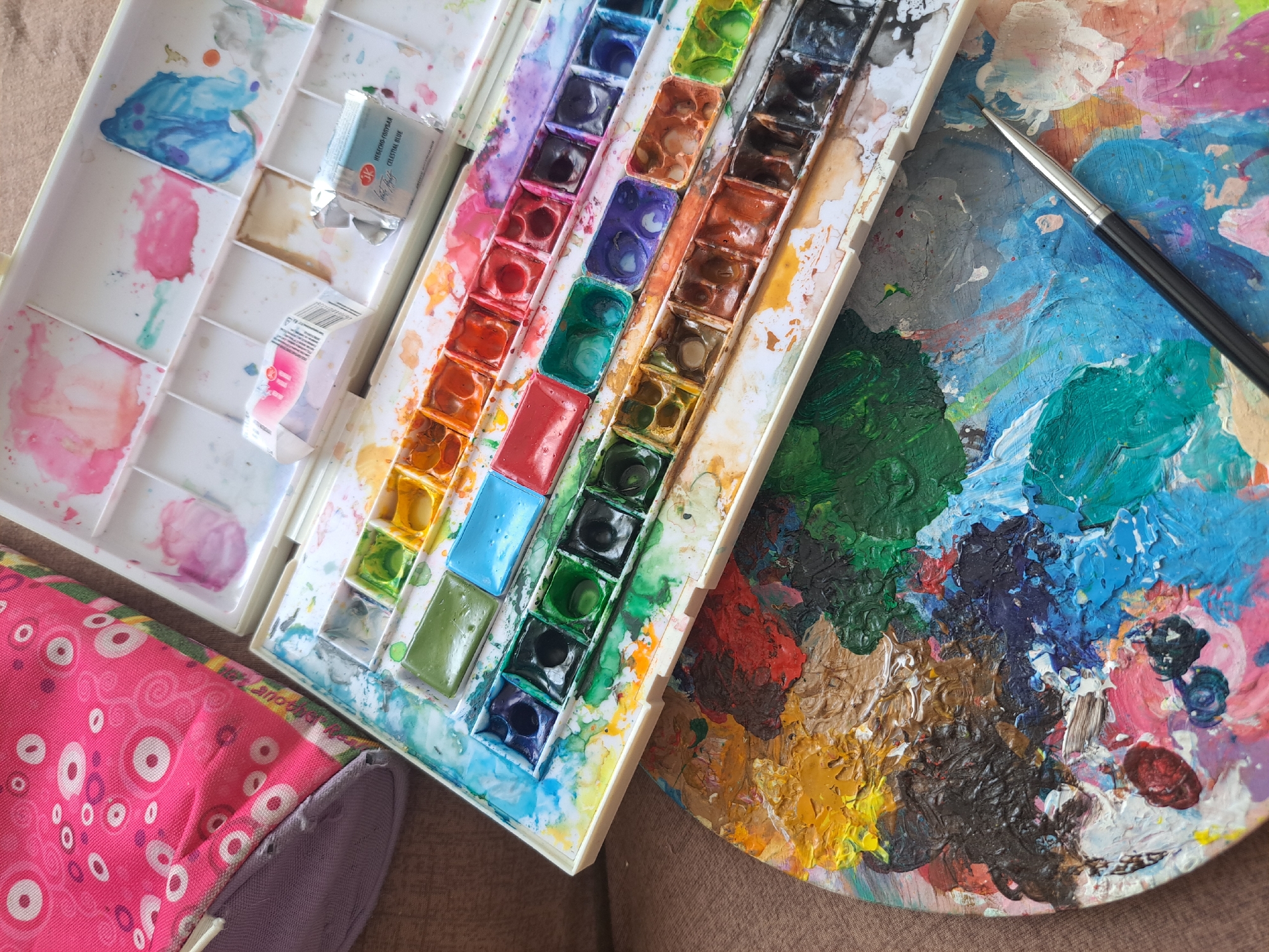 The image shows a set of watercolor paints and a palette with various colors mixed together. The watercolor set is a rectangular plastic case with multiple compartments, some of which contain dried paint cakes in different colors, while others show remnants of wet paint. Two small foil-wrapped paint tubes, one labeled "NEBESKO GORYAN CELESTIAL BLUE," are also visible within the paint case.
Next to the watercolor set is a round palette, heavily covered in thick, textured paint. The palette displays a vibrant mix of colors, including greens, blues, reds, yellows, browns, and black, suggesting an active painting session. A paintbrush with a black handle and a silver ferrule rests on the palette.
In the foreground, a corner of a pink fabric bag with a circular pattern of white and purple concentric circles is visible. The overall scene appears to be an artist's workspace, possibly in a home environment, given the casual arrangement of the art supplies. The lighting suggests it could be daytime, with natural light illuminating the scene. No specific time of day, weather conditions, or strong emotional cues are evident. The location context is Popovo, Bulgaria.