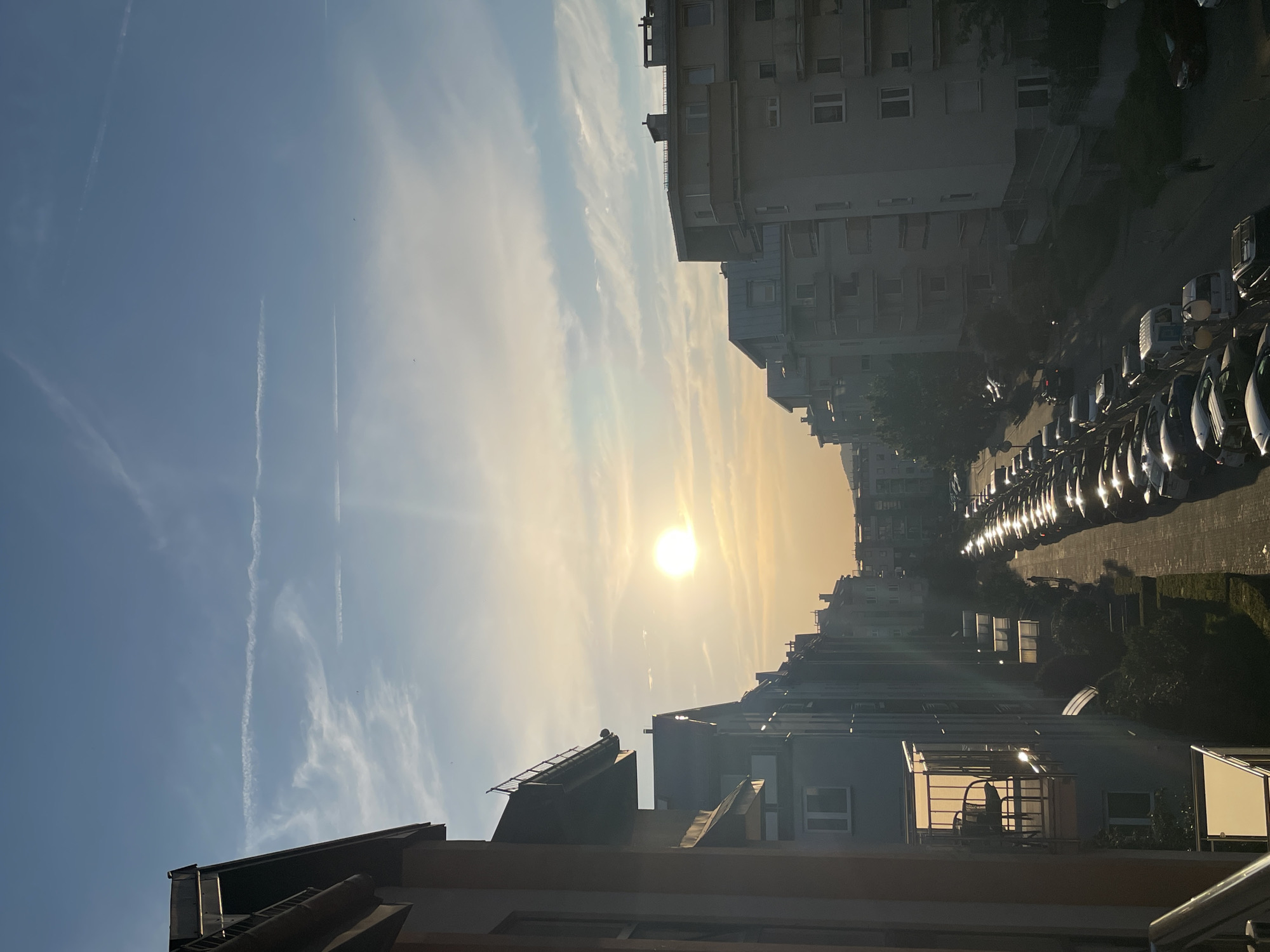 The image depicts a city street lined with apartment buildings, viewed from a slightly elevated perspective. The sun is low in the sky, casting a warm glow and creating long shadows. The sky is clear blue with wispy white clouds and several long, straight contrails from airplanes. The sun is a bright, distinct circle in the center-right of the frame.
On the street, a row of parked cars is visible, their surfaces reflecting the sunlight. The apartment buildings are modern in design, with multiple windows and balconies. Some of the buildings appear to have solar panels on their roofs. In the foreground, the roof of another building is visible, featuring angled surfaces and what appears to be ventilation or industrial equipment.
The time of day appears to be either early morning or late afternoon due to the low angle of the sun and the warm light. The weather seems to be clear and sunny. The presence of apartment buildings and parked cars suggests an urban residential setting. The overall mood is calm and serene, with the setting sun highlighting the architectural features and the sky. There is no visible text in the image.