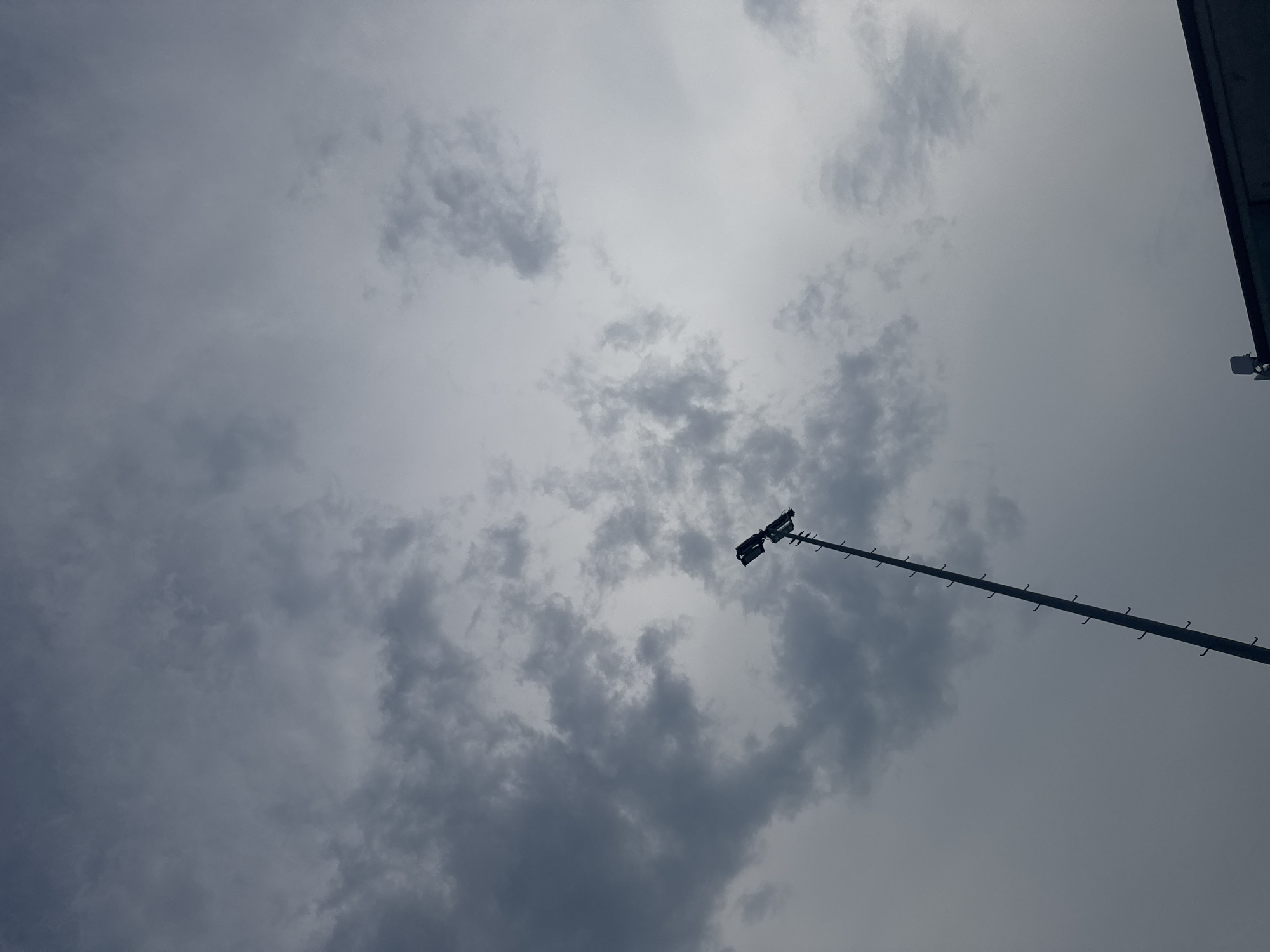 A tall, slender pole with lights at the top extends upwards against a cloudy, overcast sky. The pole is dark and appears to be made of metal, with small protrusions or steps visible along its length. The lights at the top are dark silhouettes against the bright, diffused light of the sky. The sky is filled with various shades of grey clouds, creating a somber and possibly stormy atmosphere. In the bottom right corner, a sliver of a dark, flat structure, possibly a building or roof, is visible. The image appears to be taken from ground level, looking up towards the sky. The scene is devoid of any people or animals. The overall impression is one of an outdoor urban or industrial setting under a heavy sky.
