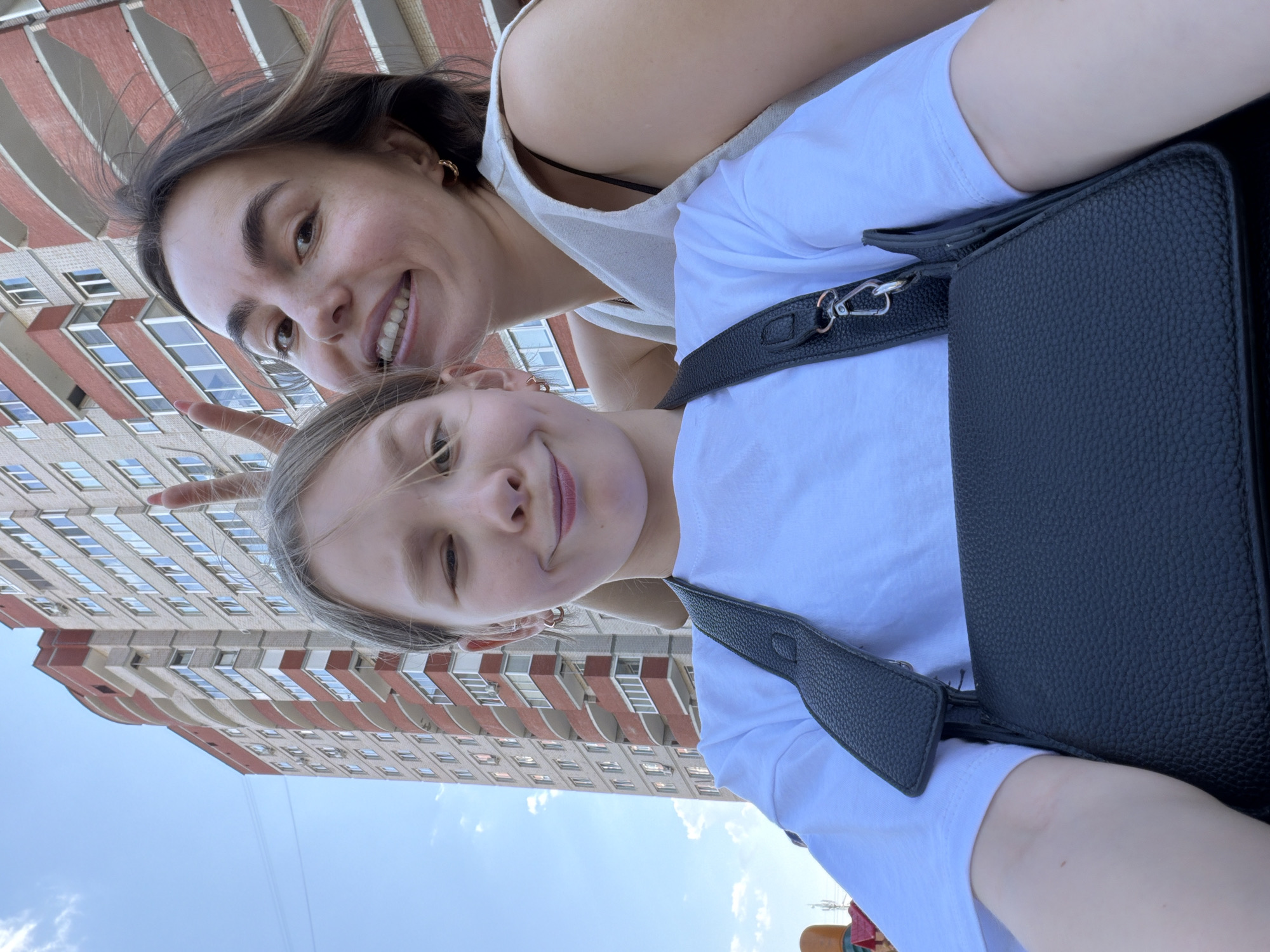 Two females pose for a selfie in Izhevsk, Russia, with a tall building visible in the background. The taller female, likely the mother, is positioned behind and slightly above the younger female, presumably her daughter. The mother smiles broadly, showing her teeth, while the daughter winks with her left eye. The mother wears a light-colored tank top, and the daughter is in a white t-shirt with a black shoulder bag. The building in the background is a high-rise structure with brick accents. The sky is visible in the upper portion of the frame.