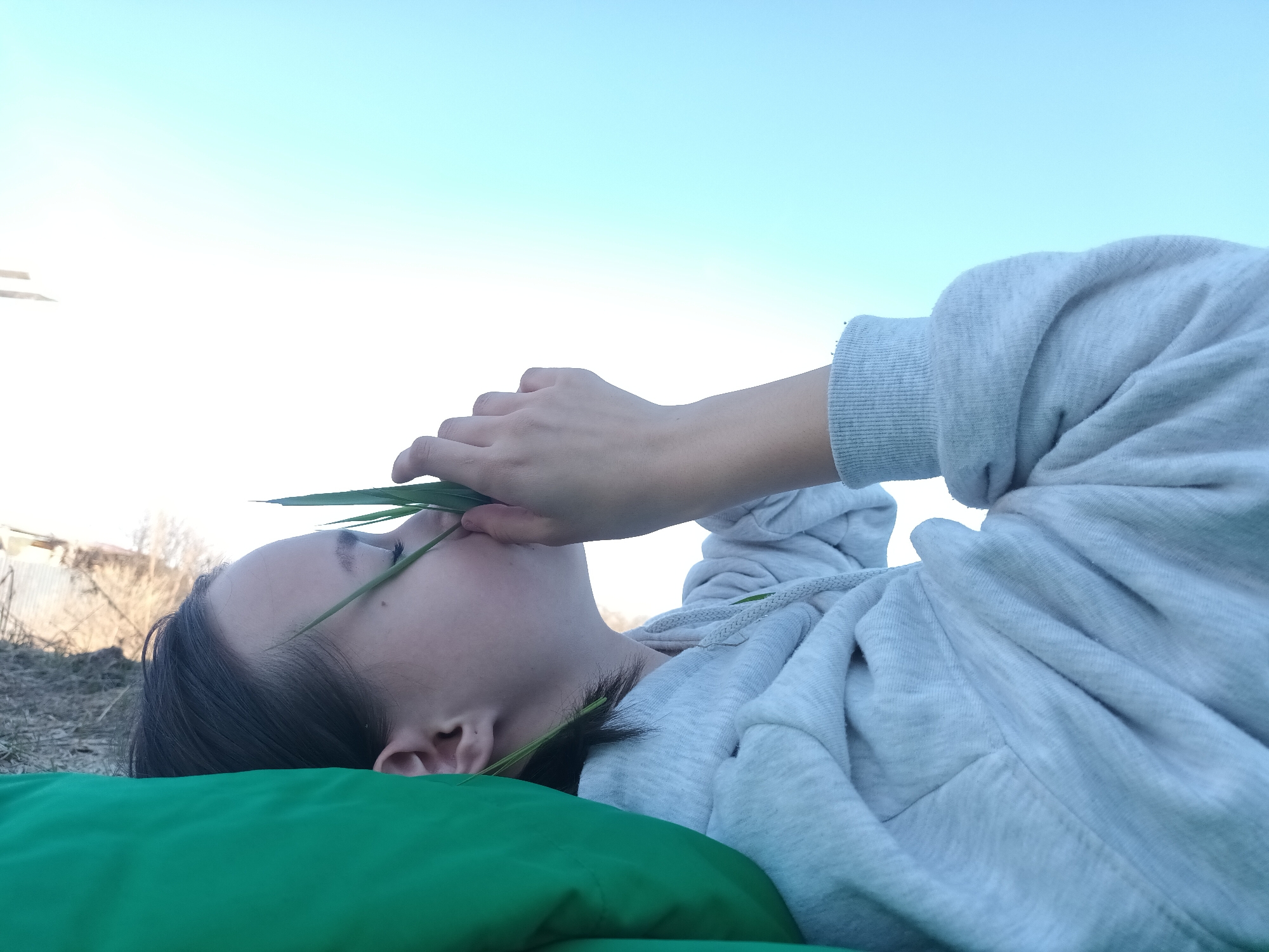 A young person with dark hair is depicted lying on their side, looking upwards, in an outdoor setting. They are wearing a light grey hooded sweatshirt. Their right hand is near their face, holding several long blades of green grass that partially obscure their nose and mouth. The person is resting on a bright green fabric or cushion.
The background features a clear, bright sky, transitioning from a light blue at the top to a nearly white hue near the horizon. In the far left background, there are hints of a dry, earthy landscape with sparse, bare trees or bushes, and some indistinct structures like a fence or distant buildings, suggesting a rural or semi-rural environment. The ground immediately around the person is not clearly visible beyond the green fabric. The overall impression is of a quiet, contemplative moment under an open sky during daylight, possibly in late winter or early spring given the bare vegetation. There is no visible text in the image.