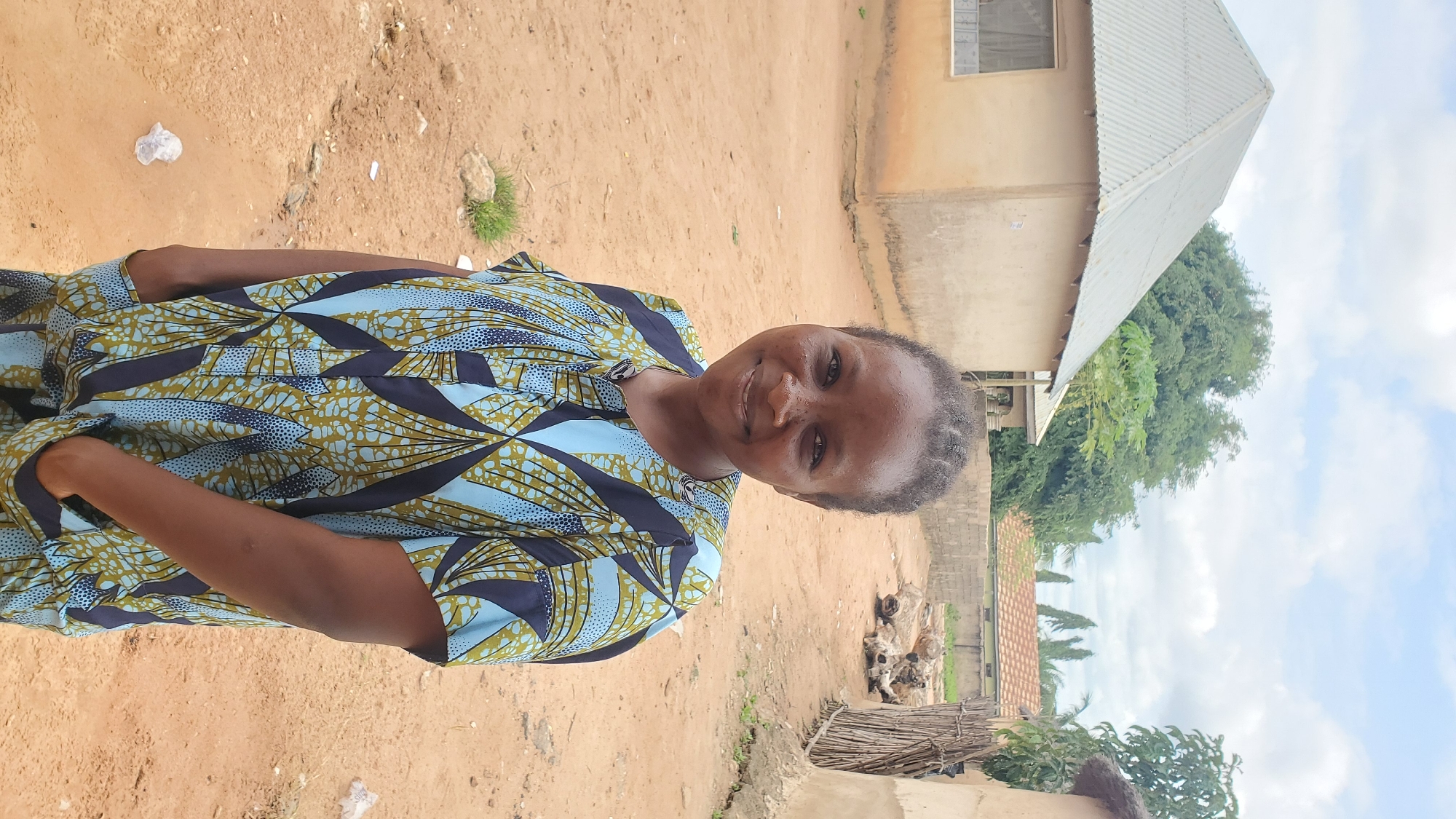 A medium shot captures a woman standing outdoors in what appears to be a rural or semi-urban area. She is smiling and looking directly at the camera. The woman is wearing a short-sleeved shirt with a blue, yellow, and dark blue geometric pattern. Her hands are in the pockets of her garment. The background consists of a dirt path, a light-colored building with a corrugated metal roof, a wooden fence, and some trees. The sky is partly cloudy. Based on visual cues consistent with images tagged as being in Jalingo, Nigeria, this scene is likely within that city.