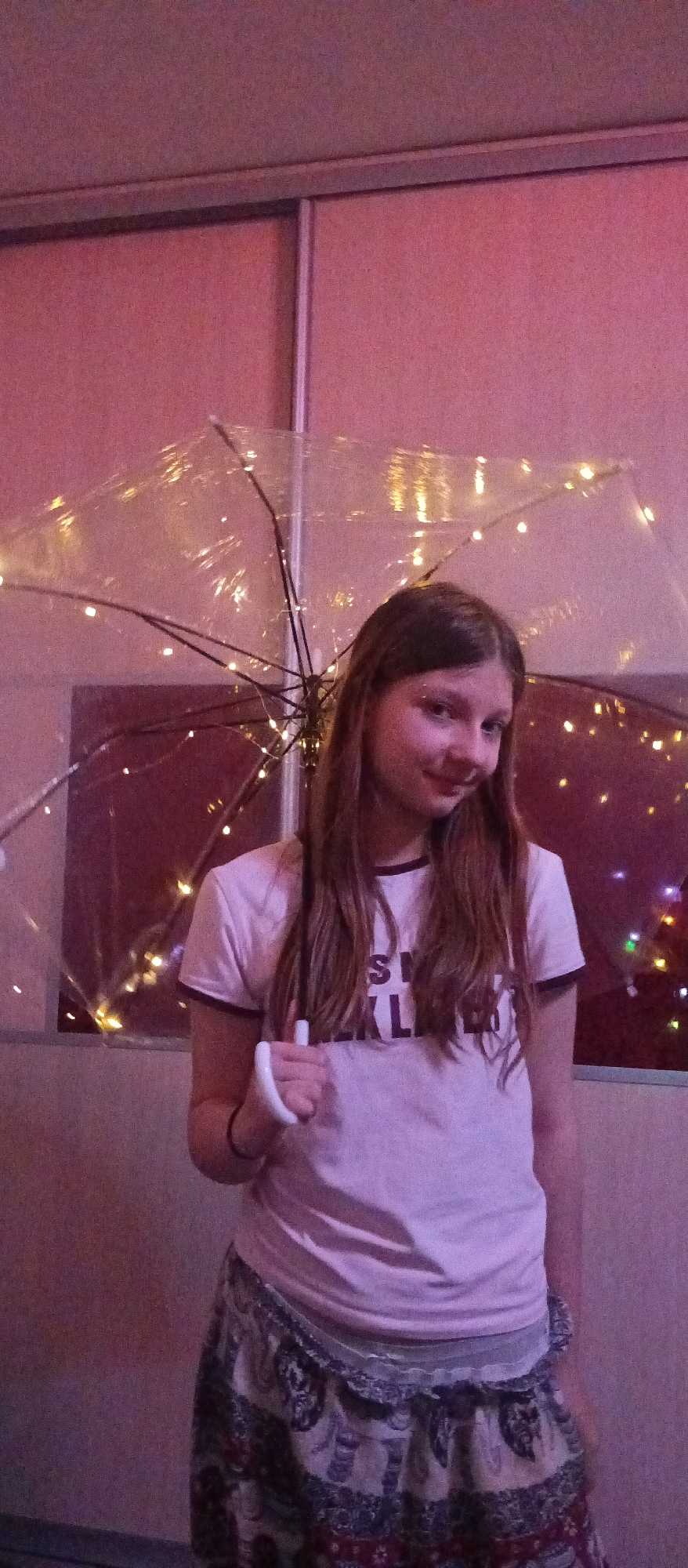 A young girl stands indoors, holding a clear umbrella adorned with string lights. She wears a light pink t-shirt and a patterned skirt. Behind her is a wall with sliding doors. The location is Unknown, Unknown.