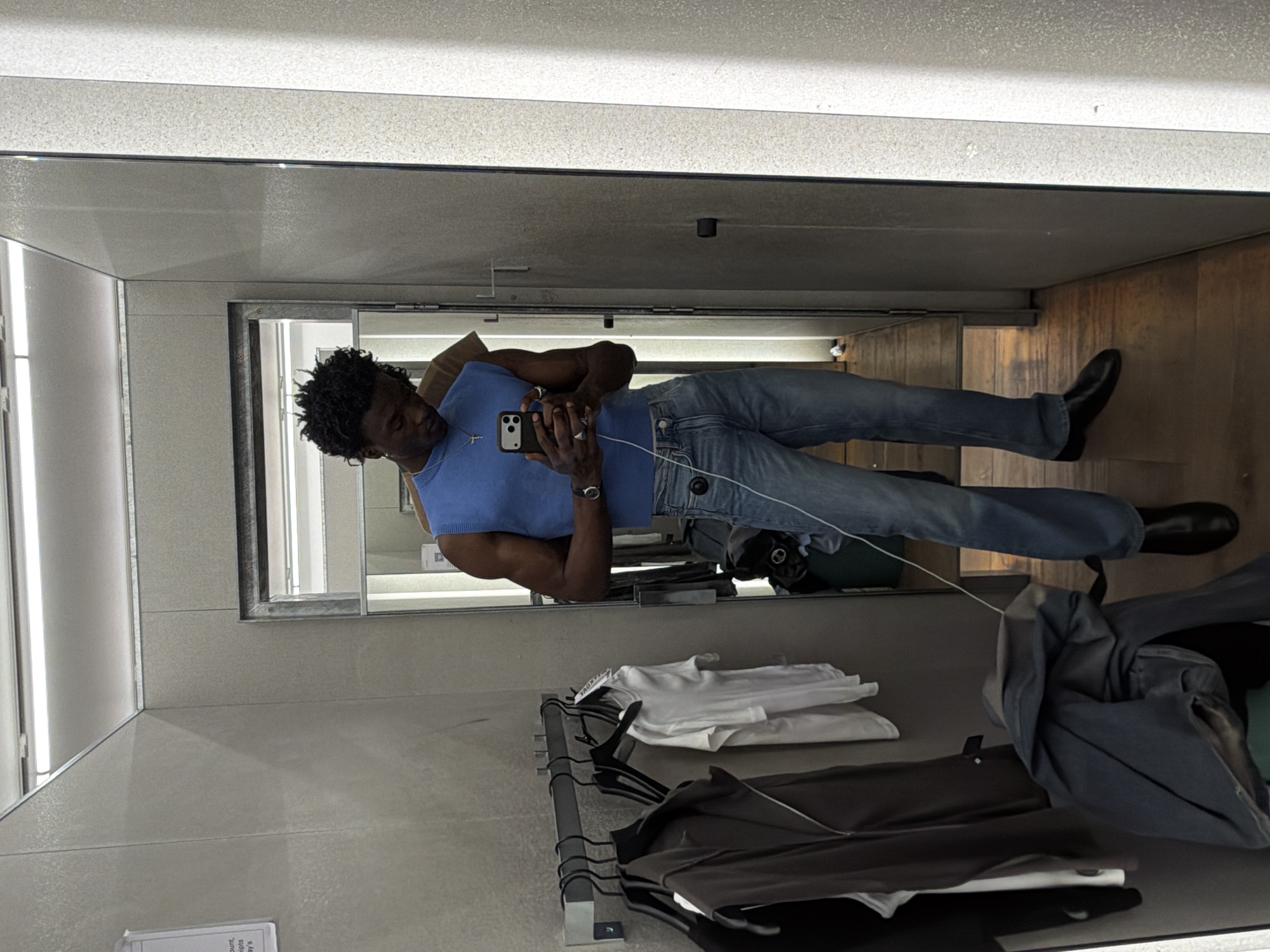 A man is depicted taking a mirror selfie in what appears to be a retail store fitting room. He is centered in the vertical mirror, holding a dark-colored smartphone with both hands to capture his reflection. The man has dark, curly hair and is wearing a blue sleeveless top, light blue denim jeans, and dark boots. A silver cross necklace is visible around his neck, and he wears a watch on his left wrist. A white charging cable is connected to his phone, dangling downwards.
The fitting room features light-colored, possibly off-white or light grey, paneled walls and ceiling, with bright overhead lighting strips reflected in the mirror. The floor is made of wooden panels. To the man's right, a clothing rack holds several folded white t-shirts on an upper shelf, while darker garments, possibly trousers or jackets, hang below. A tag on one of the hanging clothes faintly displays the brand name "ZARA." An additional piece of blue-grey clothing is loosely placed near the rack on the floor, alongside what appears to be a dark green bag. A partially visible white paper on the floor near the mirror's bottom edge contains fragmented, unreadable text, including words such as "count," "iots," and "day's." The scene conveys a moment of trying on clothing in a changing room setting.