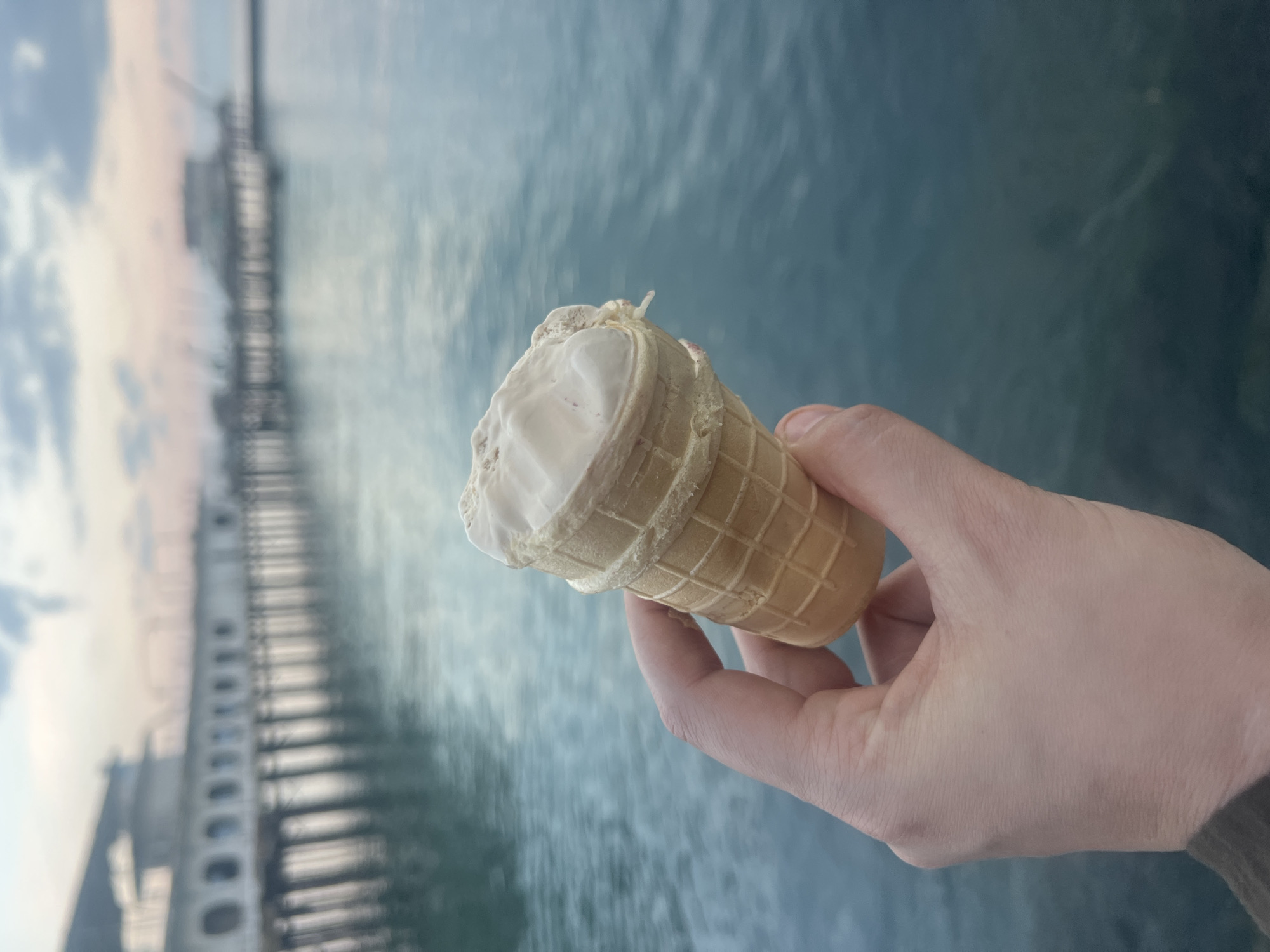A hand holds a vanilla ice cream cone against a backdrop of rippling blue water and a blurred pier. The ice cream is melting slightly, with a creamy white texture. The cone is a light golden brown with a waffle pattern. The pier, visible in the distance, appears to be made of wood and has a long, linear structure with railings. The sky above the pier is a mix of pale pink and blue, suggesting it could be dawn or dusk. The water's surface is textured with small waves, reflecting the diffused light. The overall impression is one of a serene, possibly seaside, moment, enjoyed outdoors. The image is oriented vertically, with the pier running from the top left to the bottom left.