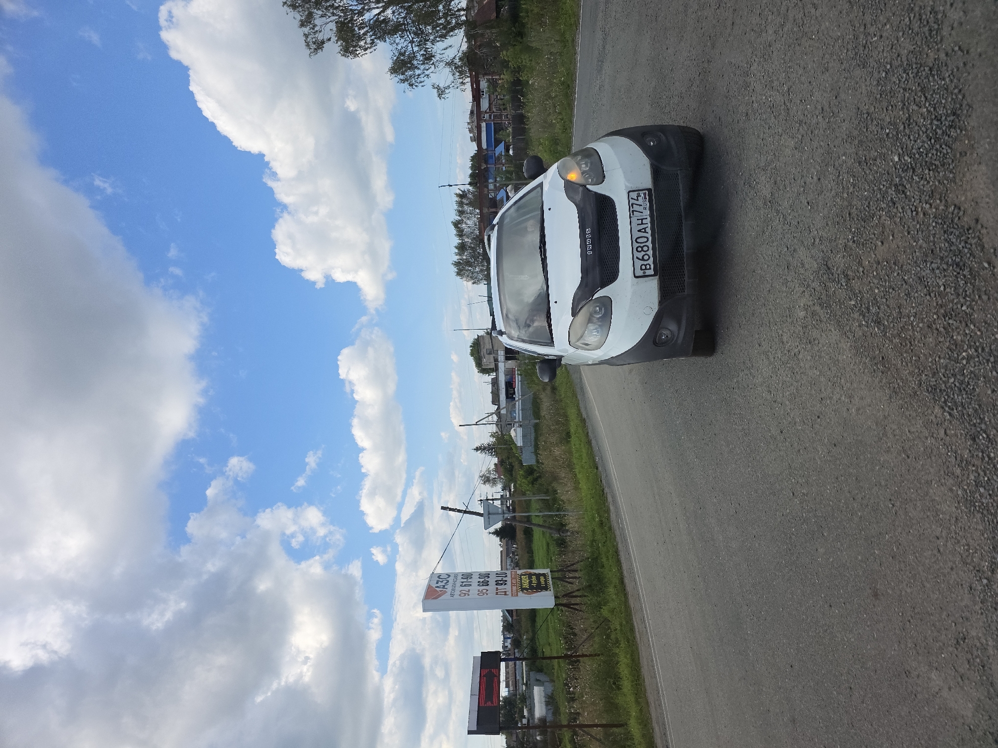 In the city of Kuluevo, Russia, on a paved road with a gravel shoulder, a white small car with a license plate reading "B680AH 774 RUS" is driving. The car has a black hood protector. To the left of the road, there is a tall white billboard with Russian text and numbers, including "АЗС," "92 61-90," "95 66-90," and "ДТ 93-10." Adjacent to the billboard is a smaller digital display showing red text. Beyond the road and billboard, there is a grassy field with scattered vegetation and trees, and in the distance, several buildings and houses are visible under a blue sky with scattered white and gray clouds.