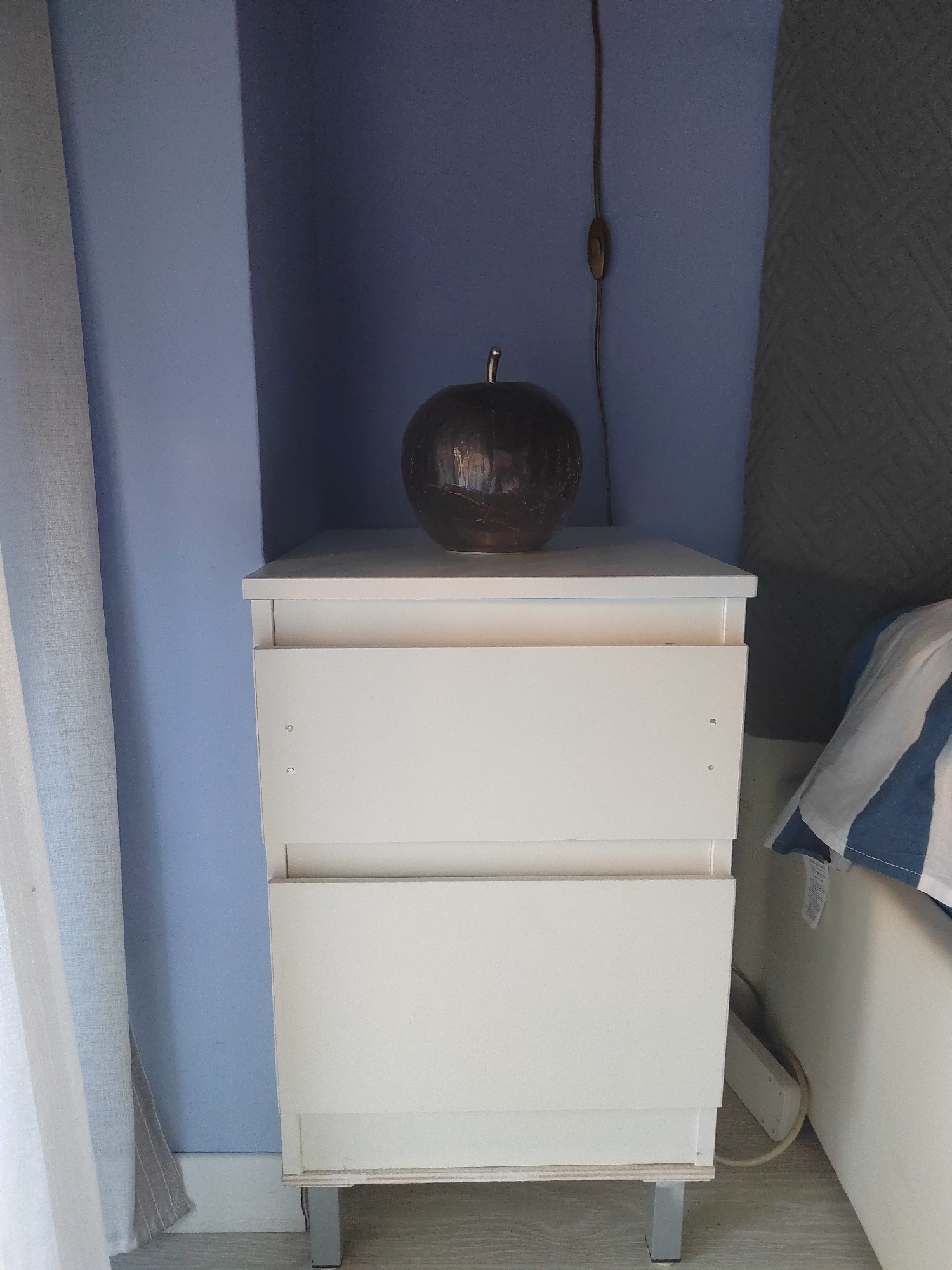 A dark brown, spherical decorative object with a metallic finish rests on top of a white two-drawer nightstand. The nightstand is positioned against a light blue wall, and to the left of the nightstand, a sheer white curtain is visible. To the right of the nightstand, the corner of a bed with blue and white striped bedding can be seen. The floor is a light wood laminate. A black electrical cord hangs down the blue wall behind the decorative object, leading to an unseen power source. The scene appears to be an indoor domestic setting.
