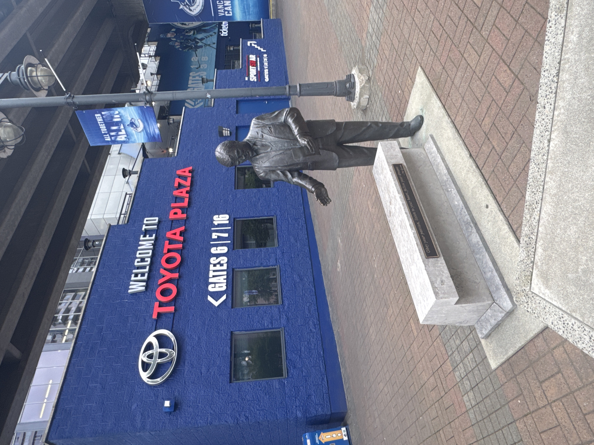 The image shows the entrance to Rogers Arena, known as Toyota Plaza, in Vancouver, Canada. A statue of a man in a suit stands on a plinth in front of the building. The building is painted blue with the words "WELCOME TO TOYOTA PLAZA" in white and red letters. Below that is the Toyota logo. To the right are the words "< GATES 6/7/16" in white. Above the entrance, there is a banner that reads "ALL TOGETHER ALL IN" with the Vancouver Canucks logo. There are also signs for "SPORT STAR GATES 1/2/3/8" and "ticket VANC CAN." The surrounding ground is paved with brick and stone. A lamp post stands next to the statue.