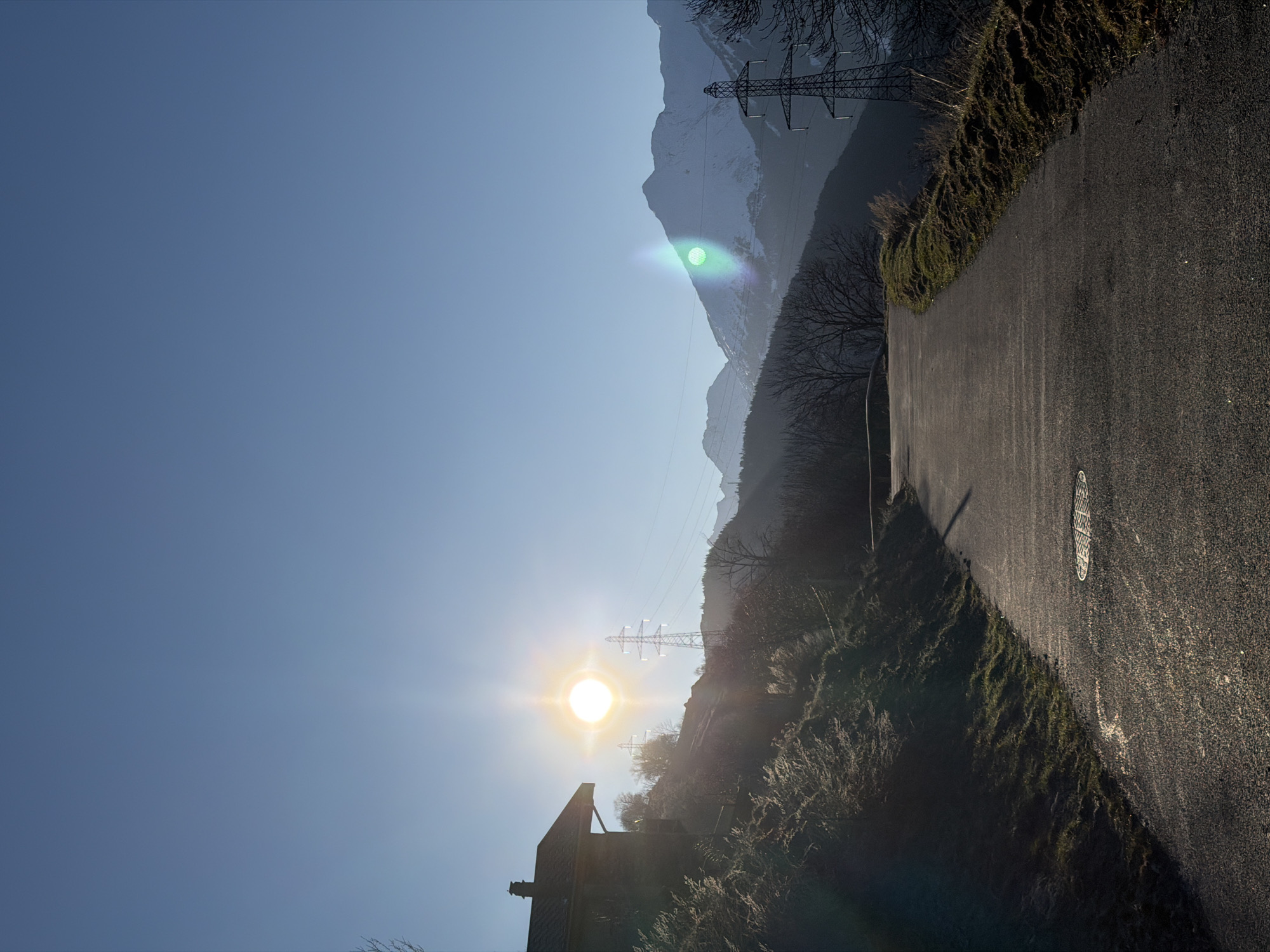 The image shows a road in Saint-André, France, with a clear blue sky partially obscured by the sun's glare. A building is partially visible in the foreground, silhouetted against the sun. Power lines and pylons stretch across the scene, traversing a mountainous landscape. The road itself is asphalt and extends into the distance. Vegetation lines the road's edge.
