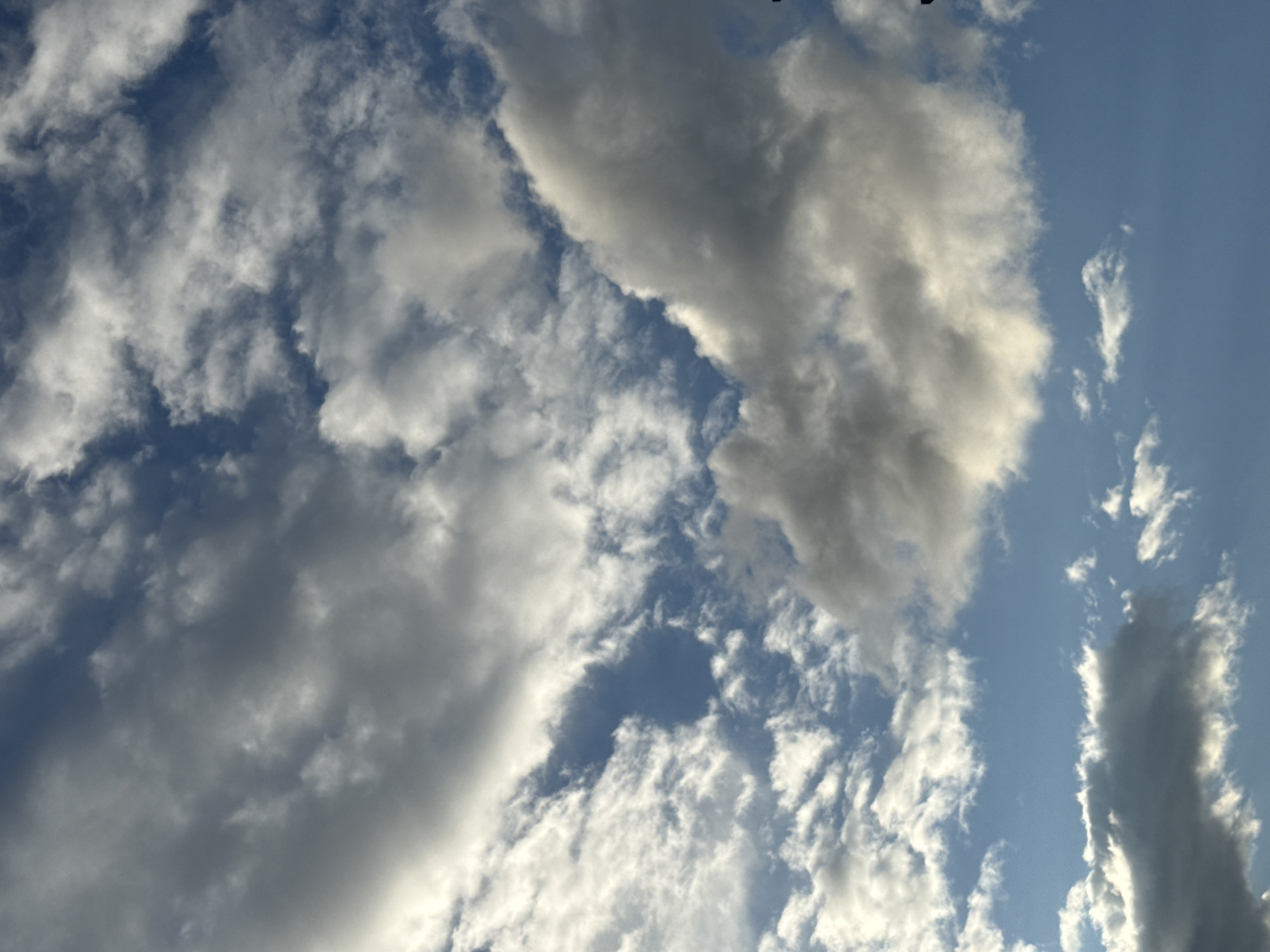The image shows a partially cloudy sky with the blue sky visible between the clouds. The clouds are white and gray, with some appearing dark and heavy. The scene is a skyview with no discernible landmarks or specific location information. There are no people, subjects, or objects visible in the image. The environment depicted is solely the sky with clouds.