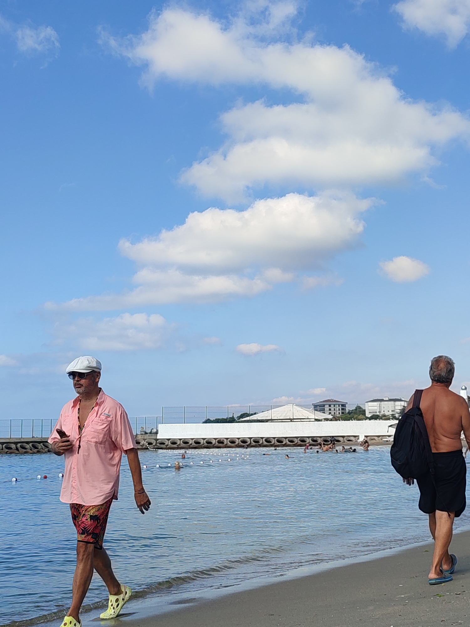 This is a vertical, eye-level shot of a beach scene on a sunny day in Kadıköy, Turkey. In the foreground, a man in a pink short-sleeved button-up shirt, patterned shorts, yellow clogs, a white cap, and sunglasses walks from left to right along the sandy shore. He is holding a smartphone in his left hand. Further to the right and slightly behind him, another man, shirtless and wearing black shorts and blue sandals, walks from right to left, carrying a black backpack over his right shoulder. In the background, the calm, blue water of the sea stretches across the frame, with a white barrier line marked by buoys in the distance. Several people are visible in the water, some swimming and others standing. Beyond the water, a white concrete structure lines the shore, and behind that, several buildings are visible, including one with a white, tent-like roof. The sky is a bright blue, dotted with fluffy white clouds.