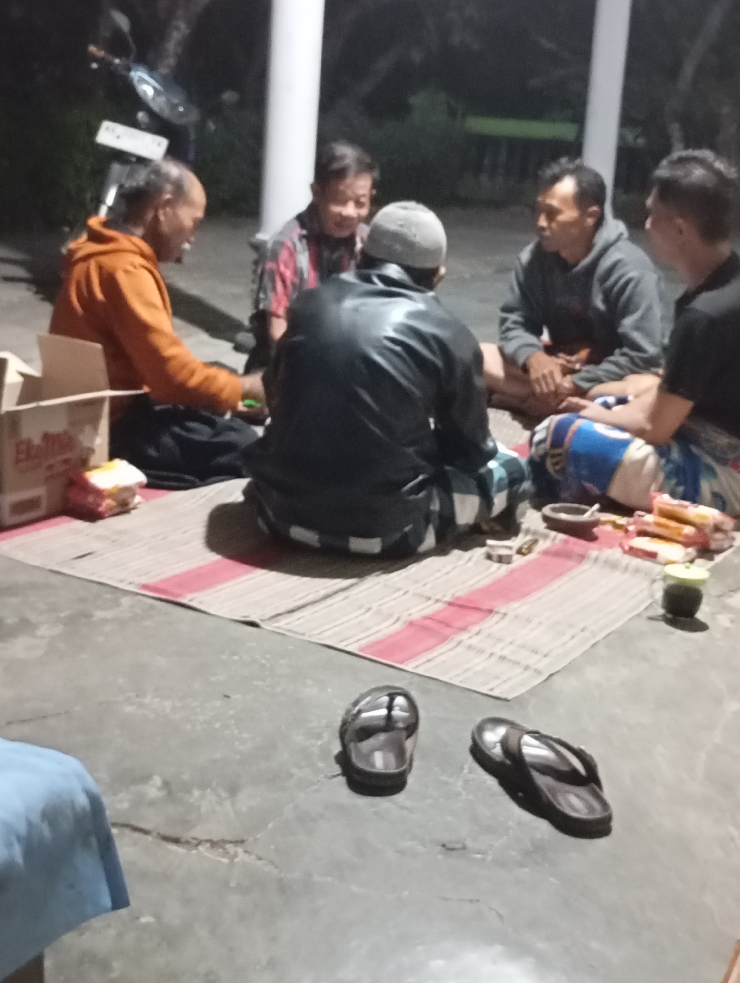 The image depicts a group of five men gathered on a woven mat outdoors, possibly in the city of Wonogondo, Indonesia, given the cultural attire and setting. The men are seated in a circle, with four visible in focus and one slightly out of frame. They appear to be engaged in conversation or a communal activity.
One man, wearing an orange hooded jacket, is positioned on the left. To his right, another man in a plaid shirt is looking towards the center. A third man, wearing a black leather jacket and a white skullcap, is seated facing away from the camera. The fourth man, in a grey hooded sweatshirt, is looking down at his hands. The fifth man, on the far right, is wearing a black t-shirt and is mostly out of frame.
On the mat, several objects are scattered. A cardboard box, possibly containing food items, is near the man in the orange jacket. There are also some packaged snacks, a mortar and pestle, and a green cup with a dark liquid. Two pairs of dark-colored flip-flops are on the concrete ground to the right of the mat.
The environment appears to be a dimly lit outdoor space with a white pillar in the background, suggesting a covered porch or patio area. The ground is concrete. The overall impression is a casual gathering of men in a relaxed setting.