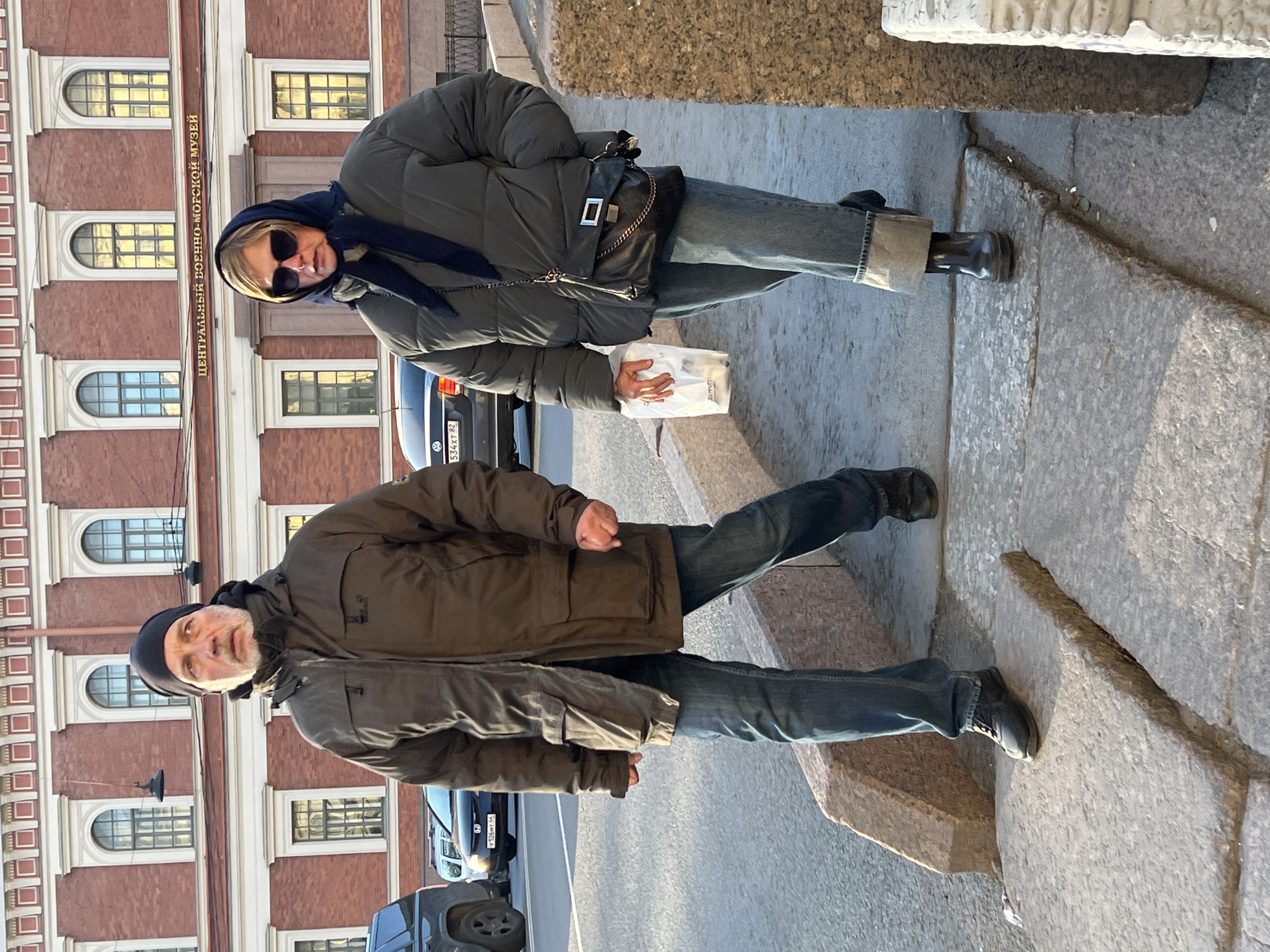 The image captures two people standing on a stone platform in Saint Petersburg, Russia, with the Central Naval Museum visible in the background. The woman, positioned in the upper portion of the frame, wears a dark green puffer jacket, jeans, and black boots. She has a navy-blue scarf wrapped around her head and is wearing sunglasses. A black purse hangs on her left side, and she holds a white paper bag in her right hand. The man, located in the lower portion of the frame, dons a brown jacket, jeans, and black shoes. He has a dark beanie cap on his head. Cars are parked on the street in the background. The scene appears to be captured during the daytime, with sunlight casting shadows on the platform and the building facade.