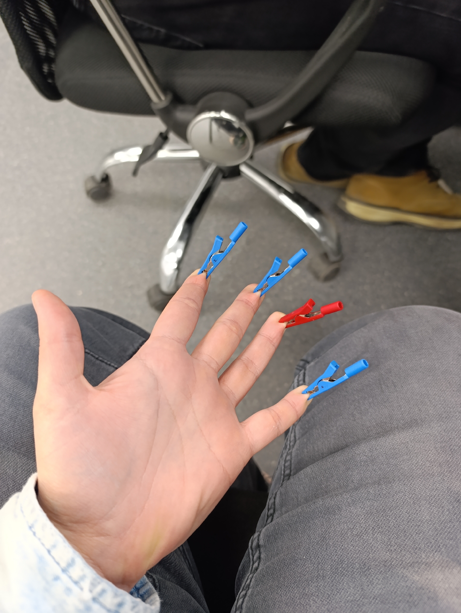 A first-person overhead shot depicts a person's hand adorned with colored electrical alligator clips on their fingernails. Three blue clips are affixed to the index, middle, and pinky fingers. A single red clip is attached to the ring finger. The person is seated, wearing grey jeans and a denim shirt. In the background, a grey office chair with a chrome base and black mesh back is partially visible. A portion of another person wearing black pants and tan shoes is also visible. The location is inside a building, most likely an office, and based on available information is not in Sofia, Bulgaria.