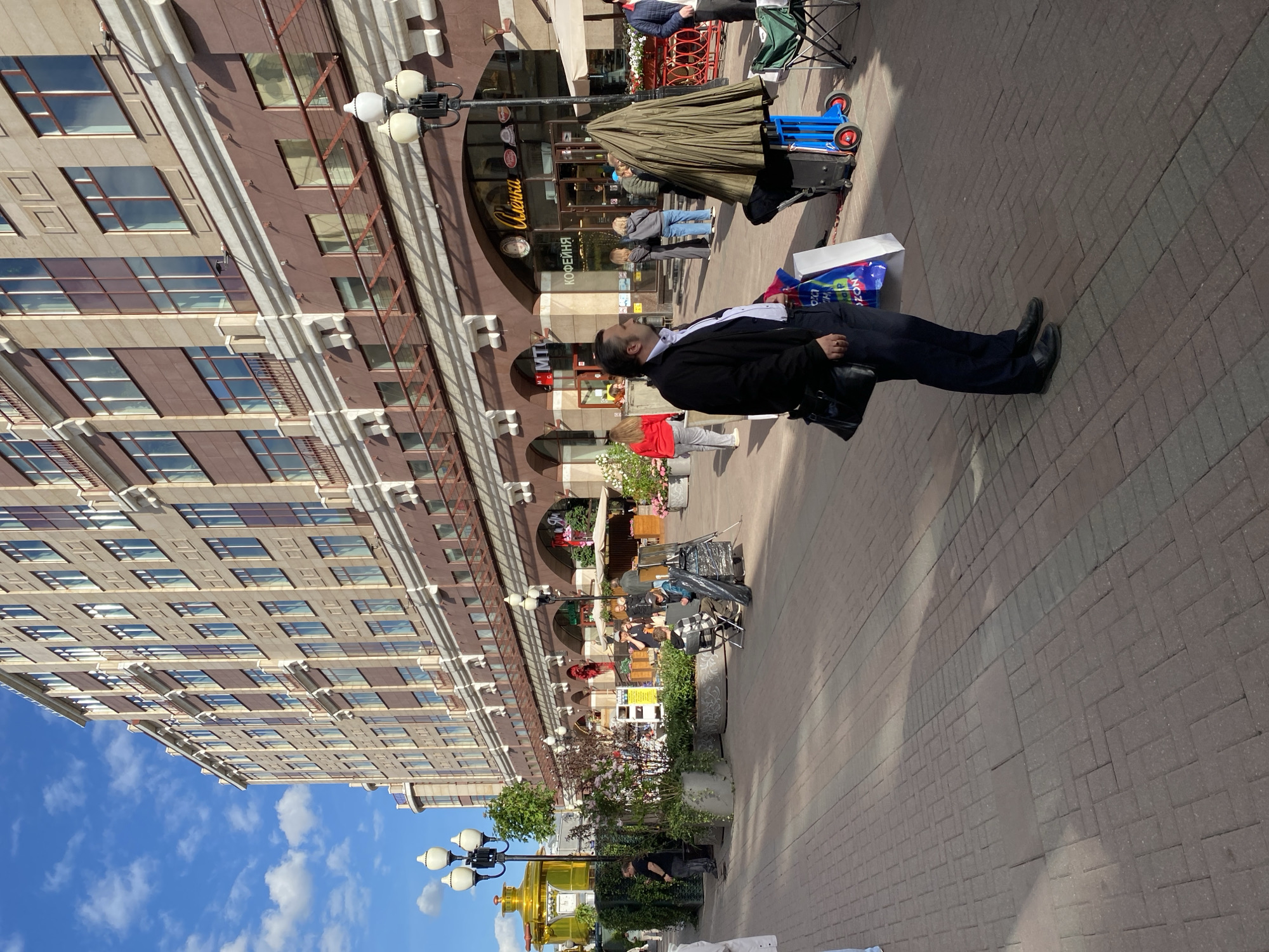 The image shows a street scene in Moscow, Russia. A multi-story building with repetitive window patterns is visible on the left side of the frame, under a blue sky with scattered clouds. Along the building's base, there is a covered walkway with shops and cafes. People are present, including a man in a dark suit and white shirt who is lying on the paved walkway with shopping bags next to him. Other people are seen walking and sitting at tables near the shops. A large golden samovar statue is visible near a street lamp. The environment appears to be a pedestrian area with brick pavement and various street furniture.