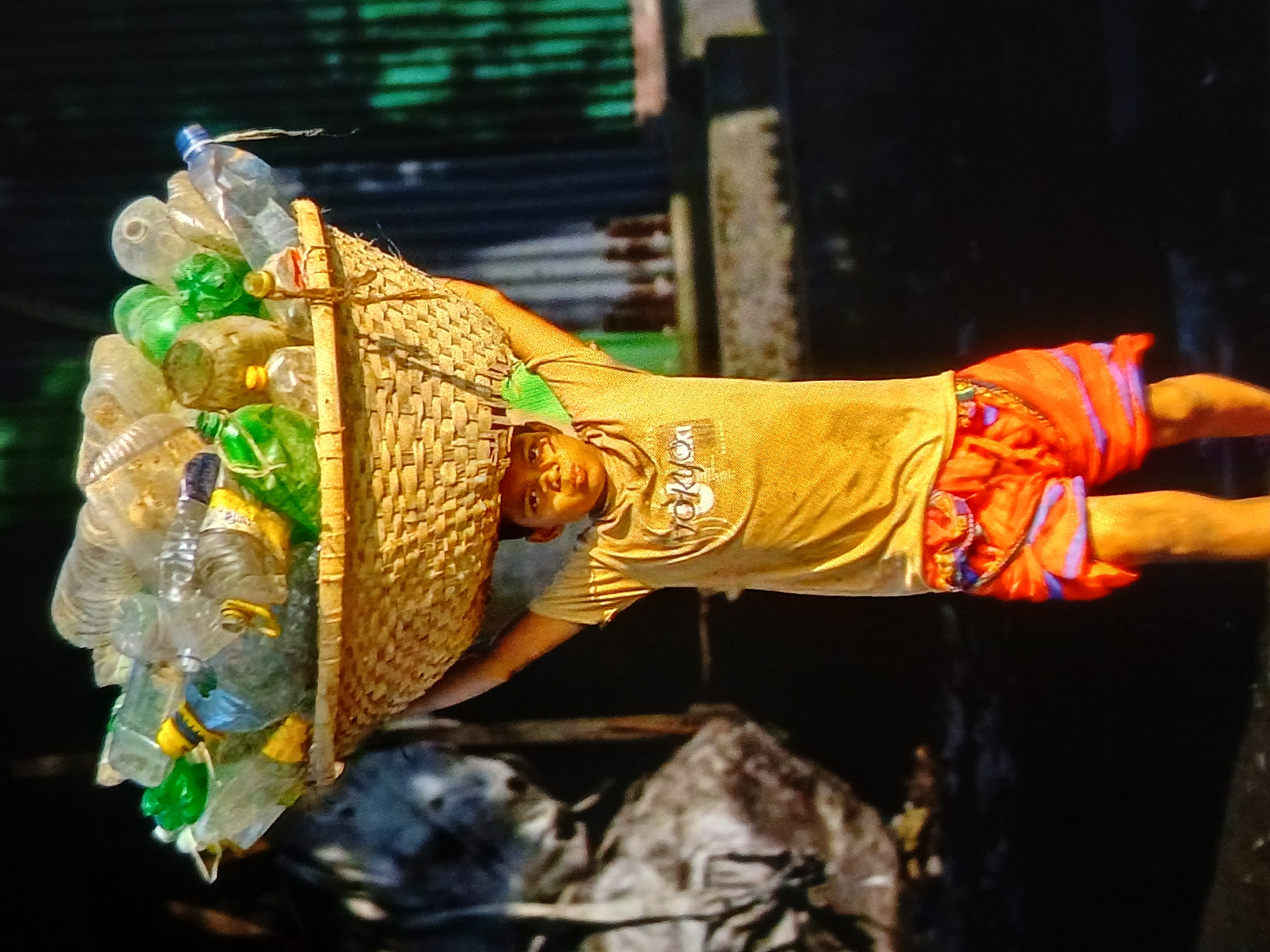 A young boy, appearing to be of Indonesian ethnicity, carries a large woven basket filled with plastic bottles on his head. He is wearing a yellow t-shirt with a graphic design and bright orange shorts. The background suggests an outdoor setting, possibly in a developing area of Setia Bakti, Indonesia, with blurred elements of what could be structures or foliage. The lighting indicates it is daytime. The boy is the central subject, actively engaged in transporting the collected bottles.