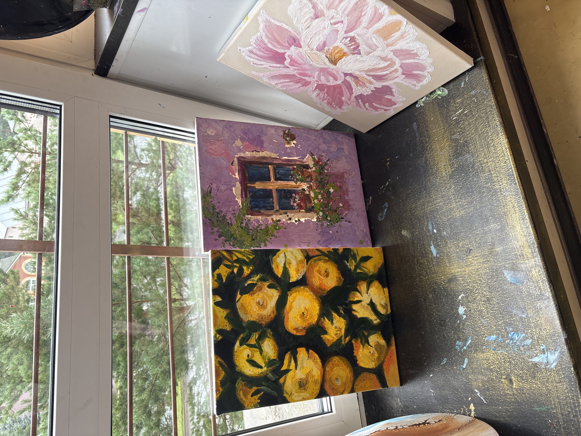 A still life arrangement of three small, unframed paintings sits on a ledge in front of a window with a view of trees and a building. The painting on the left depicts a cluster of yellow fruits, possibly lemons or oranges, against a dark background. The middle painting features a rustic window with flower boxes overflowing with pink and red blooms, set against a textured lavender wall. The painting on the right is a close-up of a large, pink flower with delicate petals. The ledge itself is dark and speckled with paint, suggesting it is a workspace. The window frame is white, and metal bars are visible through the glass. The scene is set indoors, likely in a studio or home, with the natural light coming from the window. The location is specified as Naberezhnye Chelny, Russia.