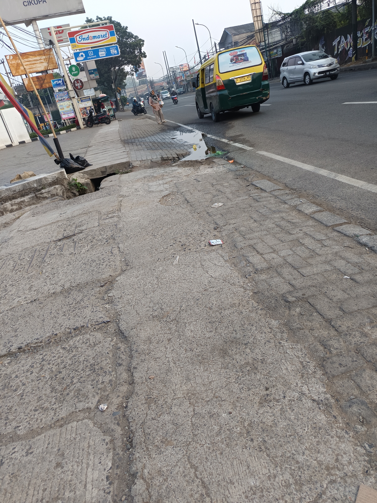 The image shows a street scene in Talaga Sari, Indonesia. A cracked and worn concrete sidewalk runs alongside a paved road. On the sidewalk, a trash bag is visible next to a drainage grate. Several signs, including one for Indomaret, are mounted on poles. A person is walking on the sidewalk, and two motorcycles are parked nearby. On the road, a yellow and green vehicle is driving, followed by a silver car. The environment appears to be a commercial or mixed-use area with buildings and trees visible in the background.