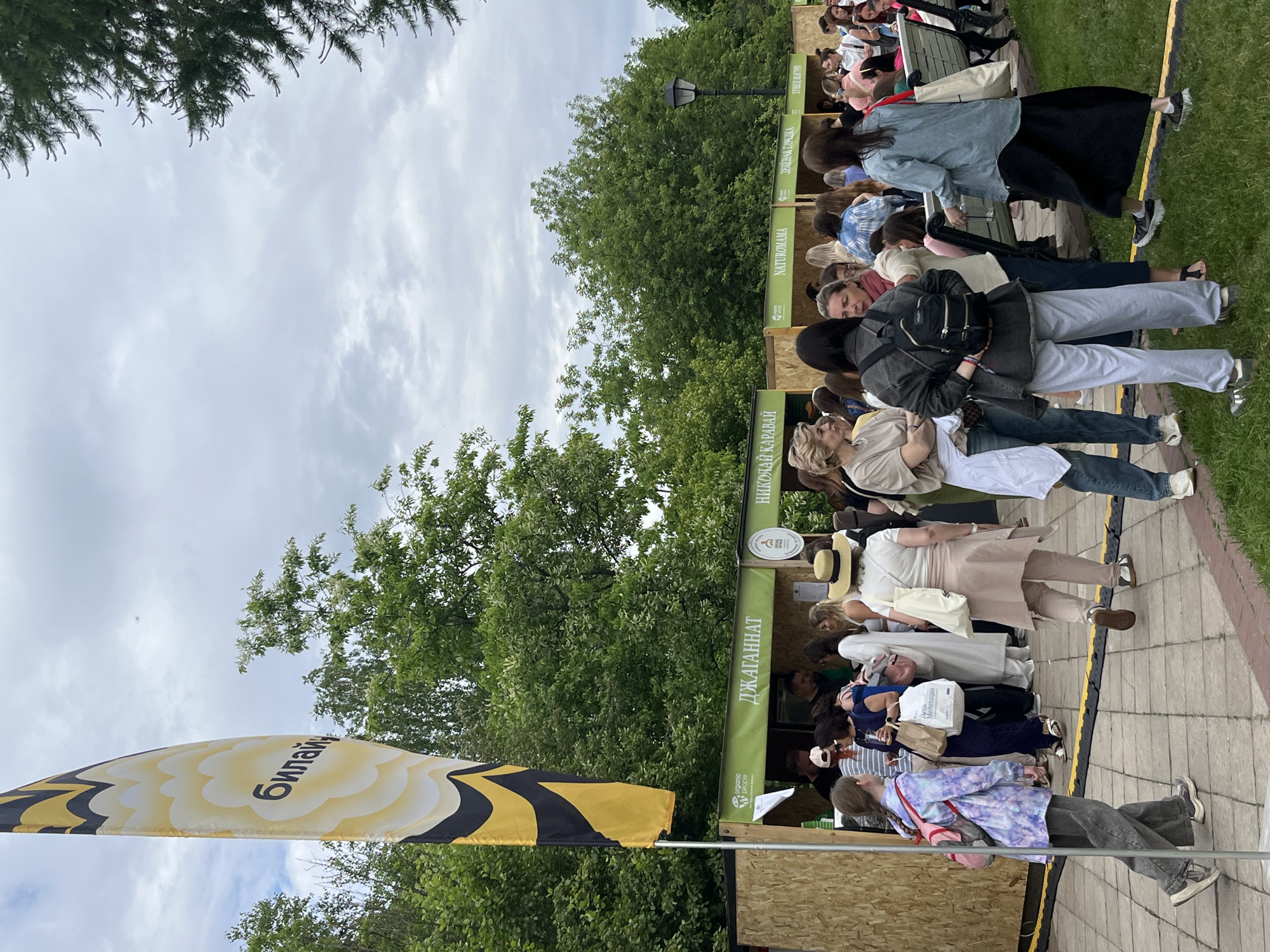 A long line of people waits at outdoor booths labeled in Russian, with trees and a cloudy sky in the background. A yellow flag with black stripes and Russian text is visible on the left. The scene appears to be in a public park or outdoor area. The location is likely in Moscow, Russia, as the booths are labeled in Russian. People stand in line, presumably to purchase something from the booths. The booths have signs, some of which include the word "Organic".