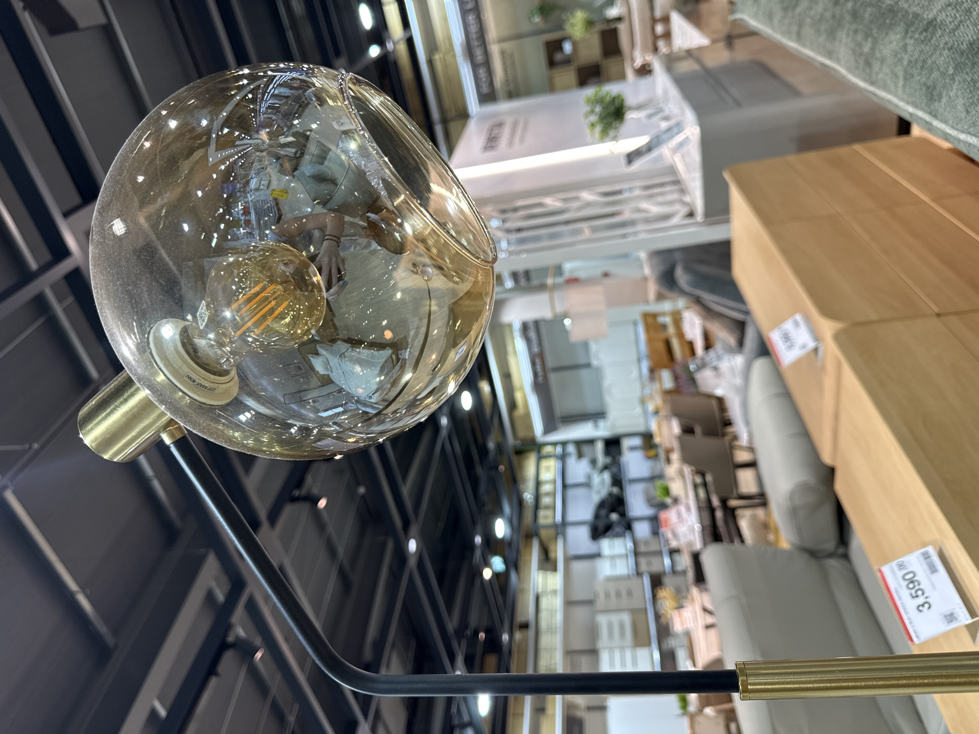 The image shows a close-up of a large, spherical, amber-colored glass lampshade with an exposed filament bulb inside, hanging from a black metal arm supported by a brass pole. The lampshade reflects the interior of what appears to be a furniture store. In the background, various furniture items are visible, including sofas, tables, and shelving units, some with price tags attached. The environment is well-lit with artificial ceiling lights. There are no people clearly visible in the foreground, but reflections in the lampshade suggest the presence of at least one person holding the camera. The location is identified as Baan Chaweng Noi, Thailand, suggesting this is likely a retail space within that area.