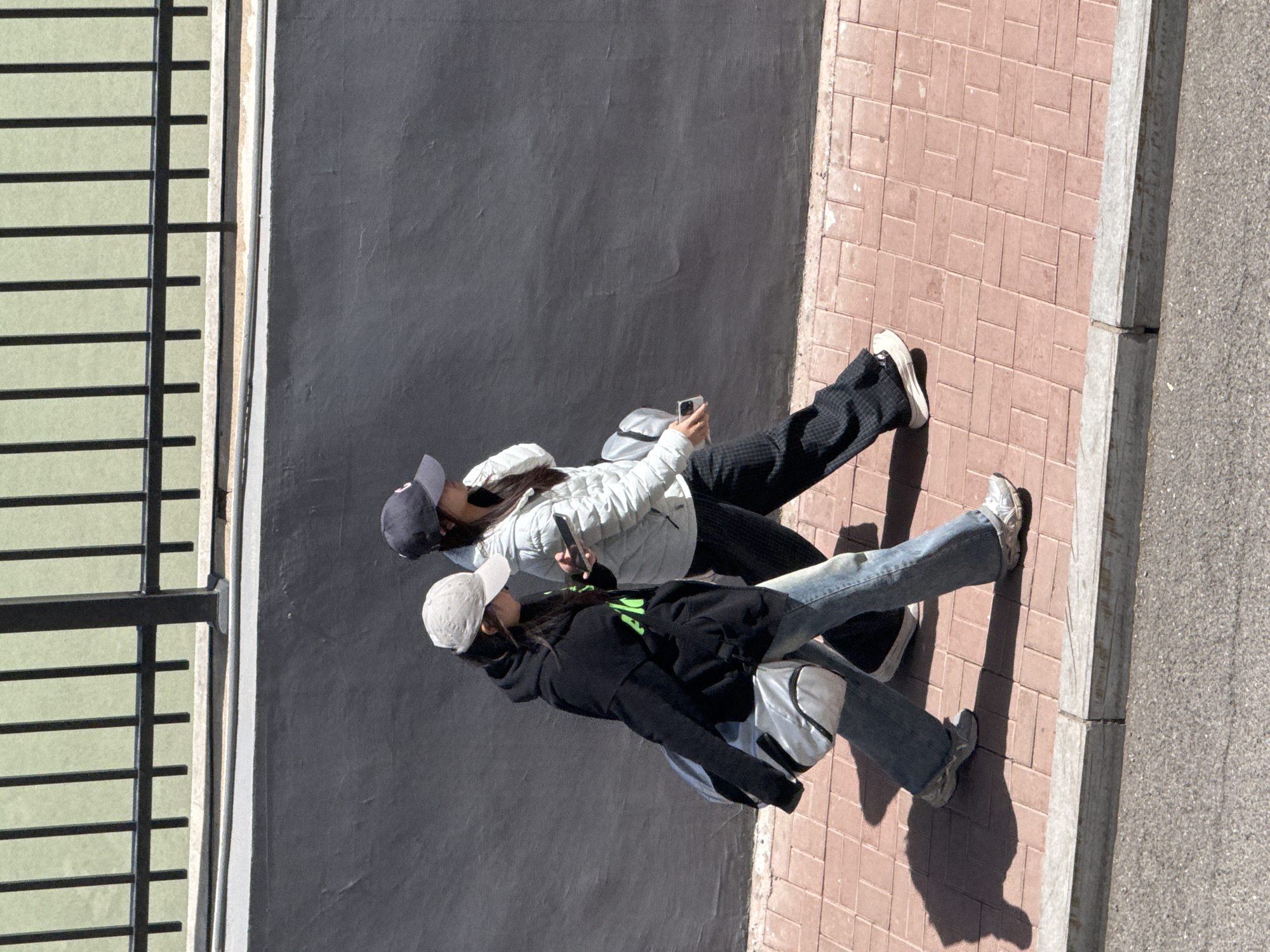 Two young women walk side-by-side on a sidewalk next to a grey wall and a metal railing. The woman closest to the curb wears a black hooded sweatshirt, blue jeans, and white sneakers, with a silver backpack. The other woman wears a light-colored jacket, dark patterned pants, and white sneakers, while looking at a phone. The sidewalk is paved with reddish-brown rectangular bricks, and the curb separates it from a gray street. The location is Zhongtun, China.