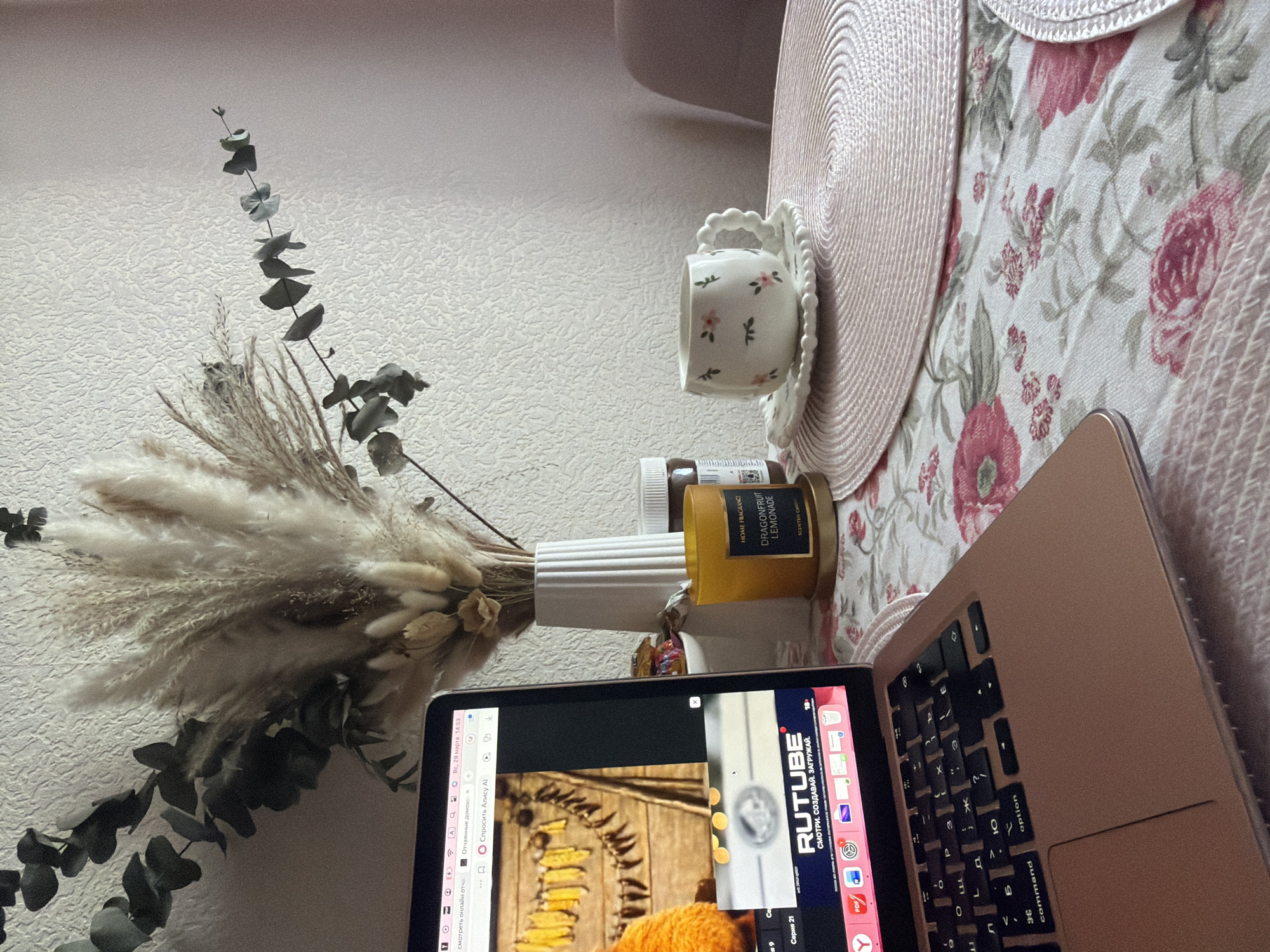 The scene is a still life composition. A bouquet of dried flowers and eucalyptus branches is arranged in a white fluted vase. A decorative teacup with a floral design sits on a saucer. A jar of scented candle labeled "DRAGONFRUIT LEMONADE" is also present. A Macbook laptop is partially visible, displaying a webpage with Russian text. The scene is situated on a floral-patterned surface, likely a bedspread or tablecloth, with a pink hat nearby. The location is not discernible from the provided image.