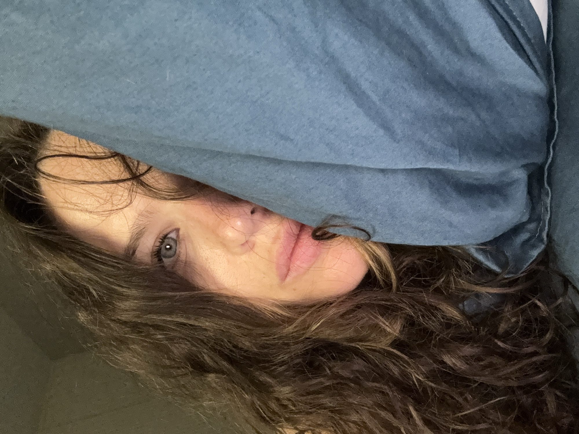A close-up image shows a person partially concealed by a blue fabric, possibly a shirt. The person's face is only partially visible, revealing one blue eye, the nose, and part of the lips. The person has wavy, brown hair that occupies a significant portion of the lower frame. The background is a dark, indistinct color. There's no information to determine the location as Dolgoprudny, Russia.