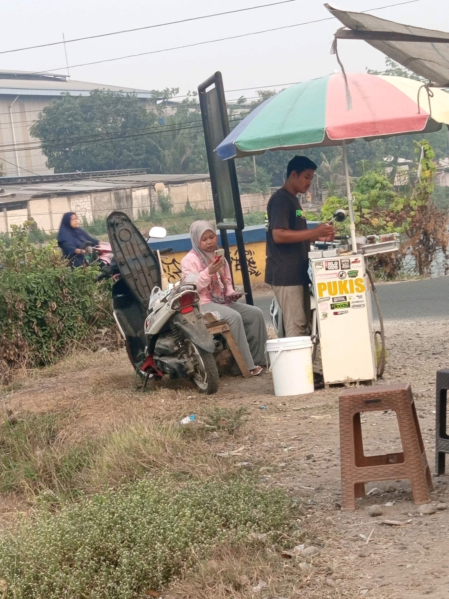 In Karawang, Indonesia, on a dirt roadside, a street vendor is preparing food under a colorful umbrella. The vendor, a man wearing a dark t-shirt and light-colored pants, stands by a cart filled with various items, possibly ingredients. In front of the cart is a white bucket. Nearby, a woman in a light pink hijab and grey pants sits on a wooden stool, looking at her phone. She is next to a parked motorcycle. In the background, a building with a light blue roof and trees are visible, suggesting an industrial or semi-urban area. The overall scene depicts a casual roadside establishment.