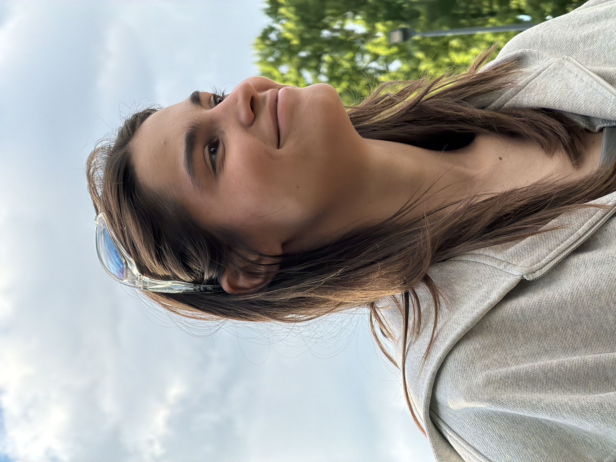 A young woman is looking upwards towards the sky. She has brown hair and is wearing a gray sweatshirt. Sunglasses are perched on her head. The background includes a cloudy sky and the green foliage of a tree. Location cannot be determined with the available information.