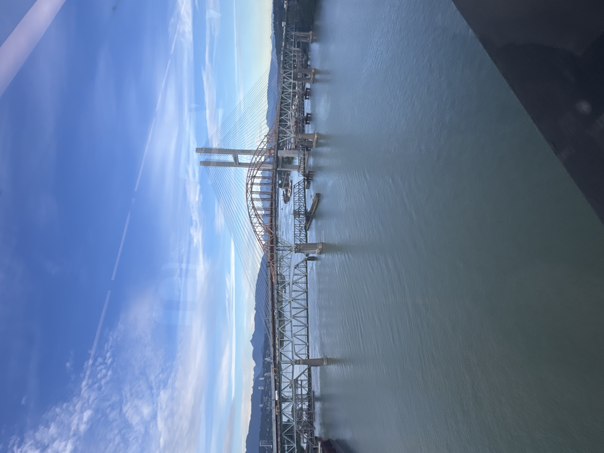 The image shows a wide-angle view of a river with multiple bridges spanning across it. In the foreground, the water is a deep blue-green, with gentle ripples on the surface. In the mid-ground, there are three prominent bridges. The closest bridge is a steel truss bridge with multiple piers supporting its structure. Behind it, another, more modern cable-stayed bridge is visible, characterized by its tall, concrete towers and suspension cables. Further in the distance, a third, older-looking bridge with a more complex structure is also present. On the far bank of the river, a city skyline with mountains in the background can be seen. The sky is a vibrant blue with scattered white clouds, suggesting it is daytime. A dark, reflective surface, likely a window, occupies the bottom right corner of the image, creating a slight obstruction. This scene is identifiable as the Fraser River in New Westminster, Canada, showcasing its vital transportation infrastructure.