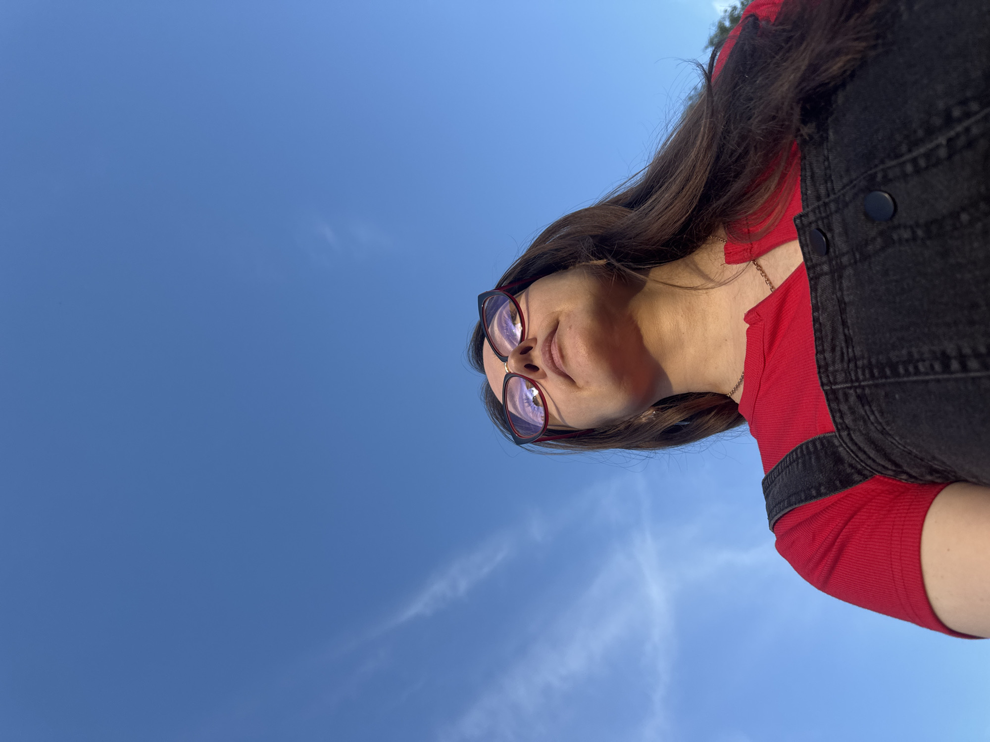 A low-angle, rotated portrait features a woman looking upwards against a bright blue sky, taken in Krasnodar, Russia. The image is oriented as if rotated clockwise, with the woman's head tilted towards the left side of the frame. She has long, dark brown hair and wears glasses with distinct red and black frames. Her facial expression is serene, with a slight, subtle smile and her mouth gently parted.
The woman is dressed in a red ribbed short-sleeved top, visible beneath the straps of dark, possibly black, denim overalls. A thin gold chain is faintly visible around her neck. The lighting suggests it is daytime, with direct sunlight illuminating her face, indicating a clear and sunny day. The background consists entirely of a vast, clear blue sky, with only a few faint, wispy white clouds discernible. No specific landmarks or other objects are visible to indicate a precise location within Krasnodar, aside from the sky itself. There is no visible text in the image.