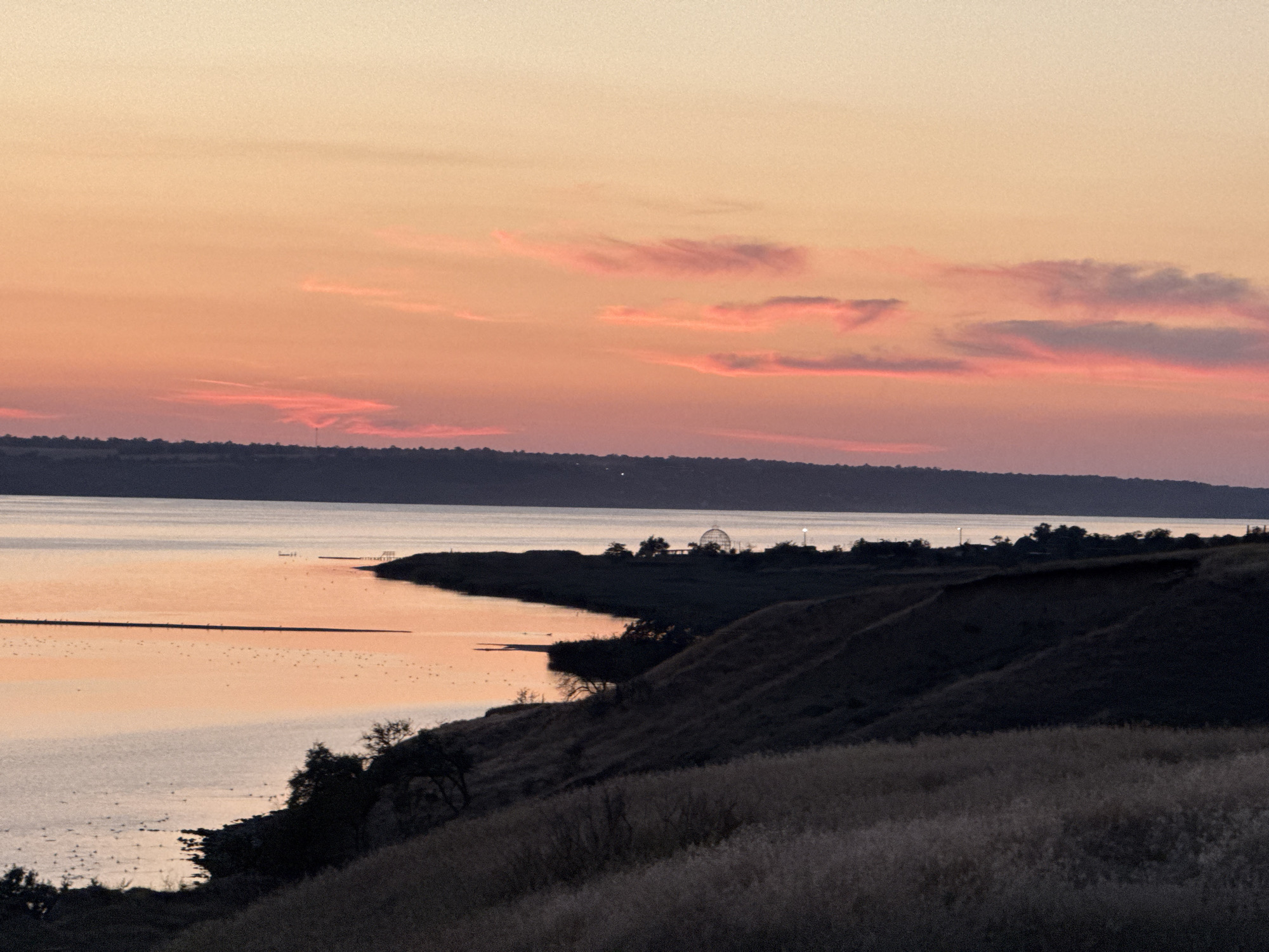 An outdoor, horizontal, medium shot captures a sunset over a body of water, likely a reservoir or large river, near Protopopivka, Ukraine. The sky is painted with hues of pale yellow, peach, and pink, with wispy clouds reflecting the sunset colors. In the middle ground, a dark, undulating shoreline is visible, with a long, thin structure stretching into the water and a domed building with a thin spire on the far shore, possibly a greenhouse or observation deck. The water itself reflects the warm tones of the sky. In the foreground, a sloping hill is covered in dry, golden-brown grass, with silhouetted trees dotting the landscape. The scene evokes a sense of natural beauty and tranquility.