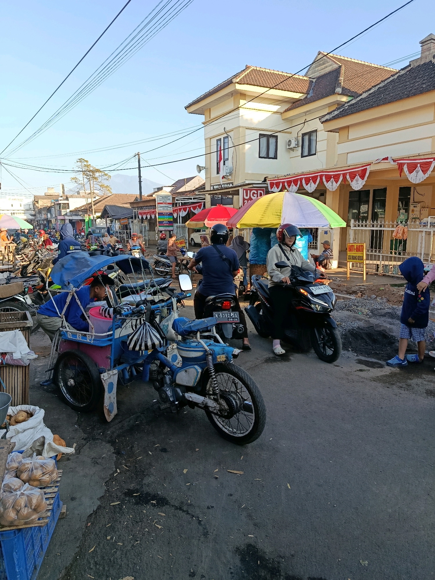 The image depicts a bustling street scene, likely a market or roadside area, in Bululawang, Indonesia. The foreground is dominated by a blue and pink motorized tricycle, with a person in a blue shirt attending to it. To the right of the tricycle, a woman on a black motorcycle wears a helmet and a light-colored jacket. Behind her, another motorcycle is parked, with a person in a dark blue shirt seated on it. Further into the background, a street lined with various buildings and shops is visible, with several parked motorcycles, people walking, and colorful umbrellas providing shade. The buildings have tile roofs and yellow and cream-colored facades. A street sign with "HATI-HATI ADA ORANG KERJA" (Caution, workers are present) is posted on the right side of the road. The ground is paved and appears somewhat worn, with patches of dark debris. A young child in a blue hooded jacket and blue checkered shorts stands on the right, looking downwards. The overall atmosphere suggests a lively commercial environment under a clear sky with visible power lines overhead.